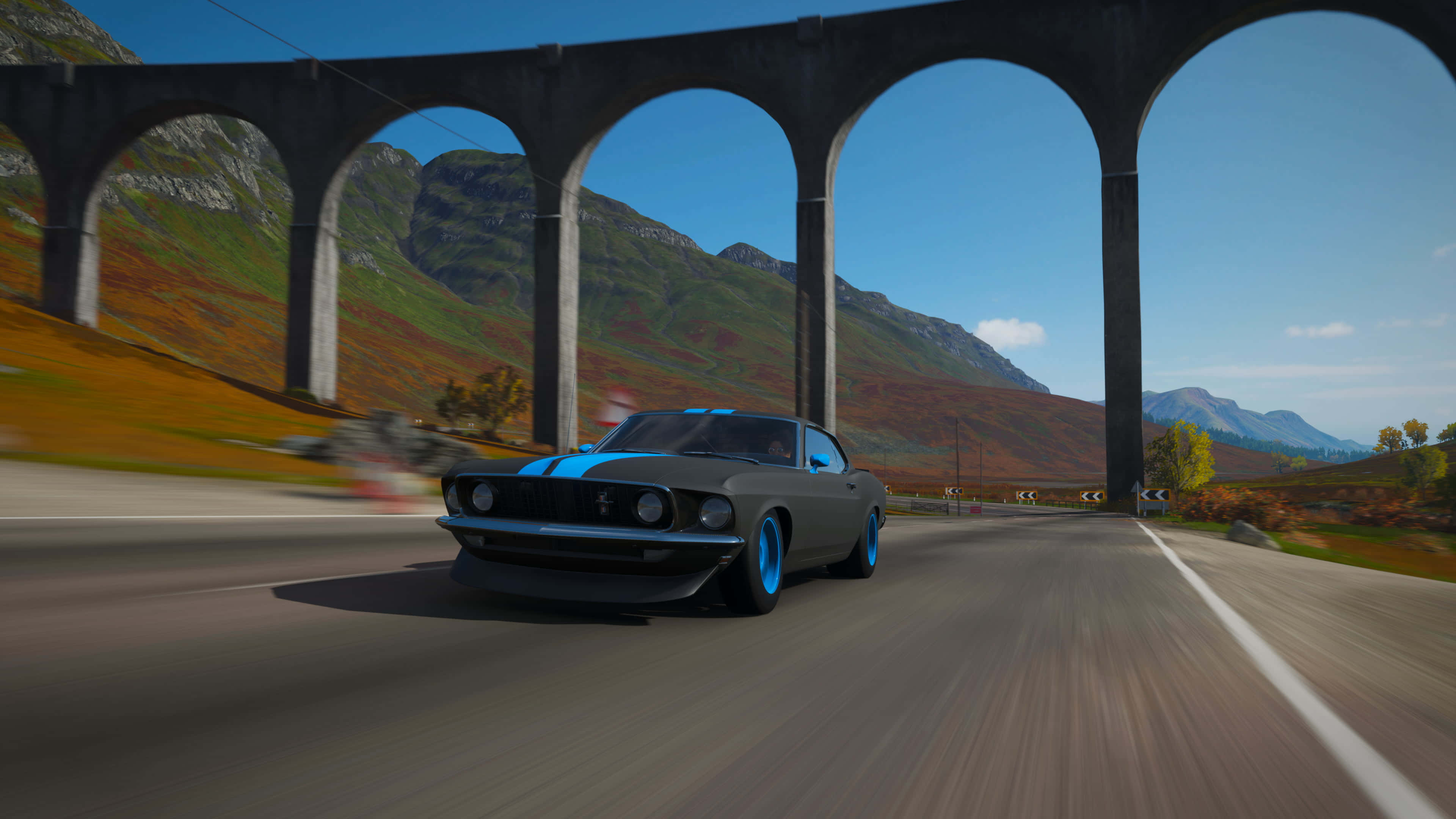 Forza Horizon 5 4K 桌布 壁紙 — 一輛帶有藍色點綴的時尚灰色肌肉車在石頭高架橋下穿過多山的秋季景觀飛馳。