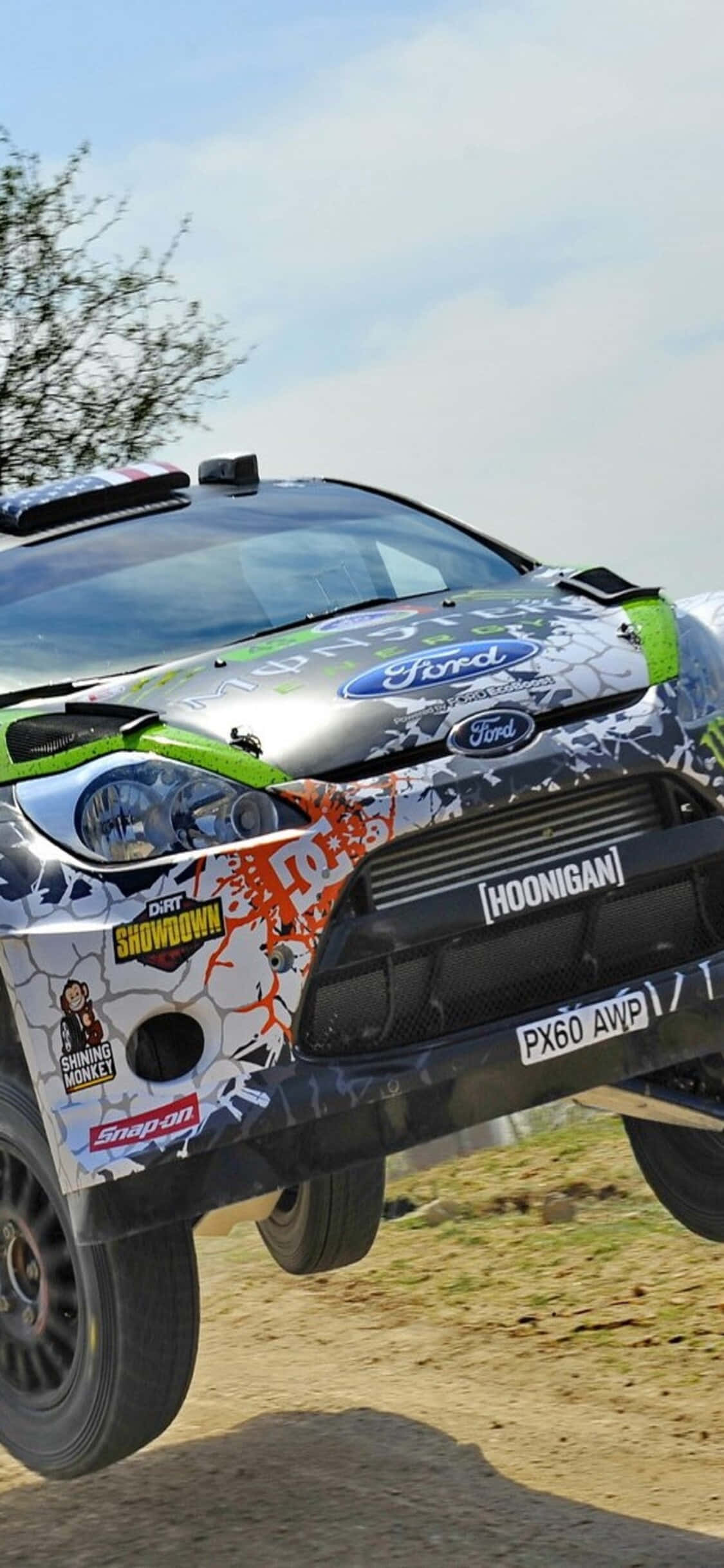 iPhone Xs Dirt Showdown 桌布 壁紙 — 一輛帶有醒目圖案的福特拉力賽車在明亮的天空下飛躍泥地賽道。