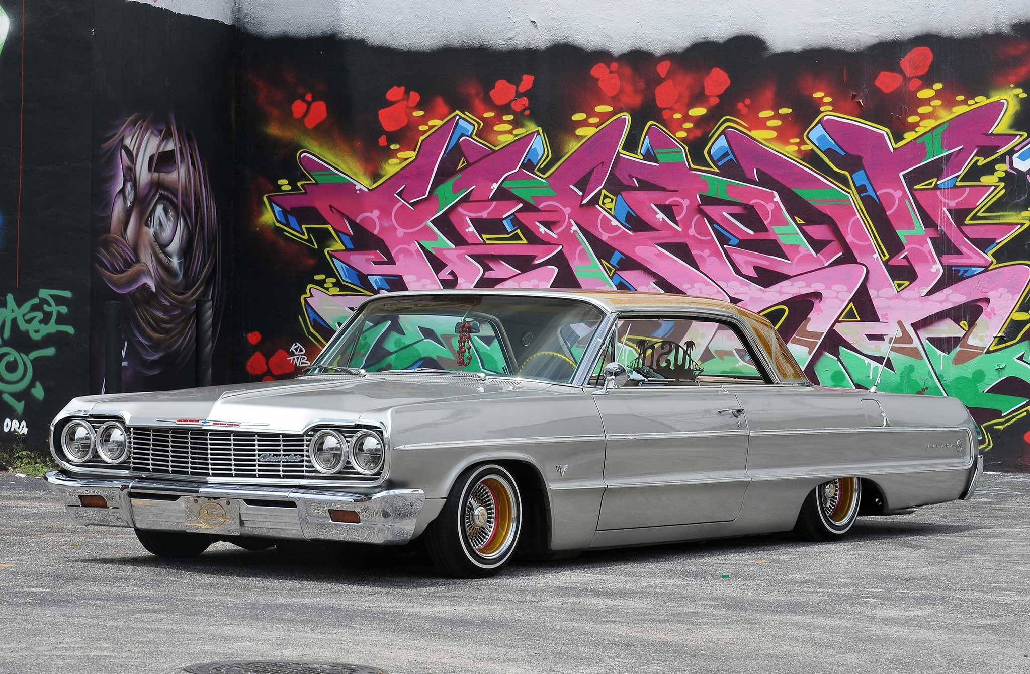 Lowrider 桌布 壁紙 — 一輛帶有金色飾邊的銀色經典低底盤汽車停在充滿活力的塗鴉牆前。