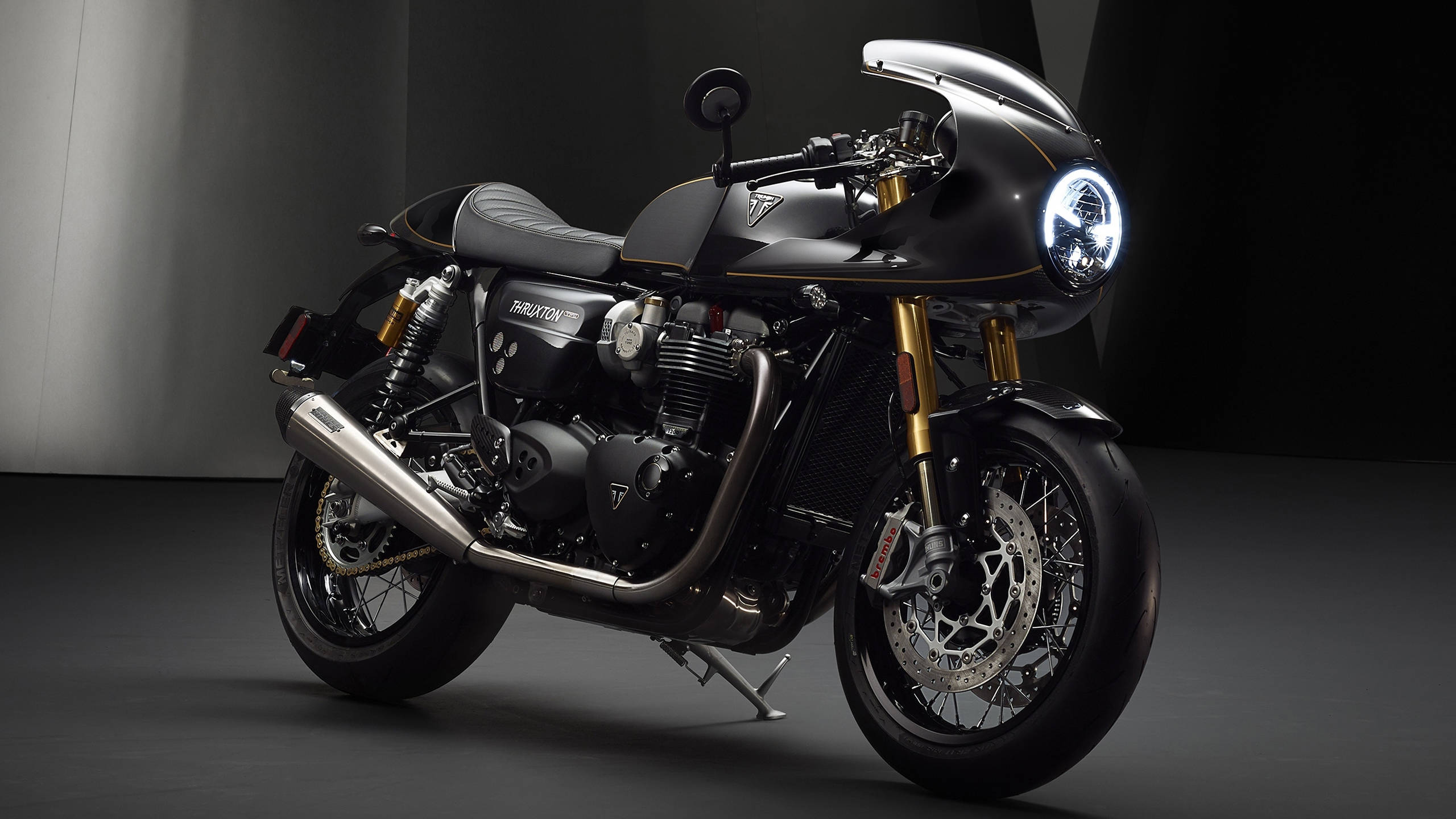 Triumph 桌布 壁紙 — 一輛帶有金色點綴和發光車頭燈的時尚黑色 Triumph 摩托車，在簡約的攝影棚環境中蓄勢待發。