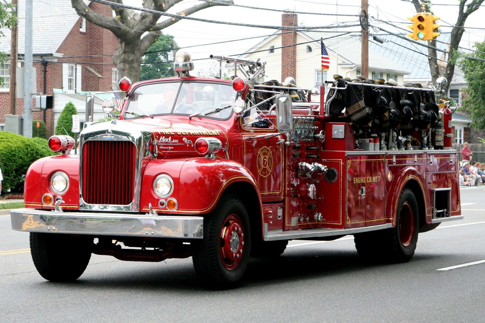 Fire Truck 桌布 壁紙 — 一輛帶有鍍鉻細節和金色飾邊的復古紅色消防車行駛在兩旁種滿房屋和樹木的郊區街道上。