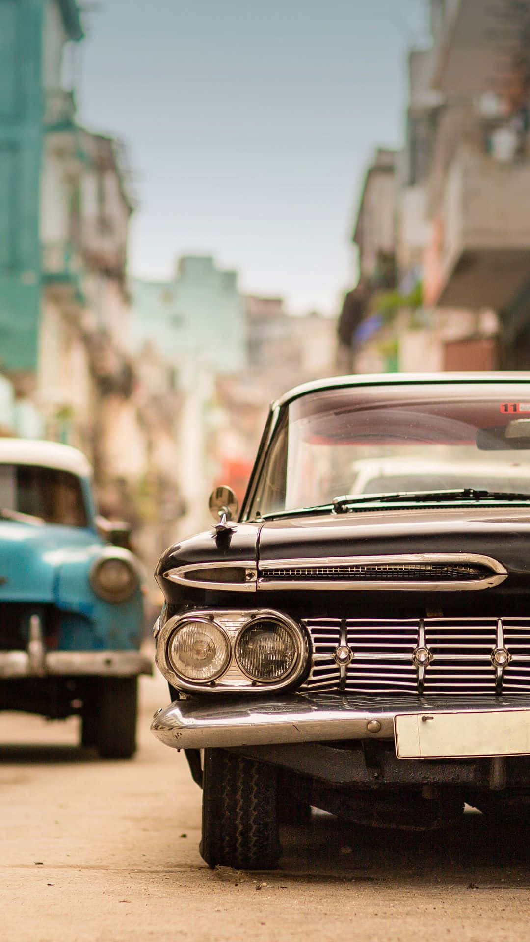 Vintage Car iPhone 桌布 壁紙 — 一輛帶有鍍鉻細節的復古黑色汽車停放在陽光明媚的城市街道上，街道兩旁是色彩繽紛的殖民時期建築。