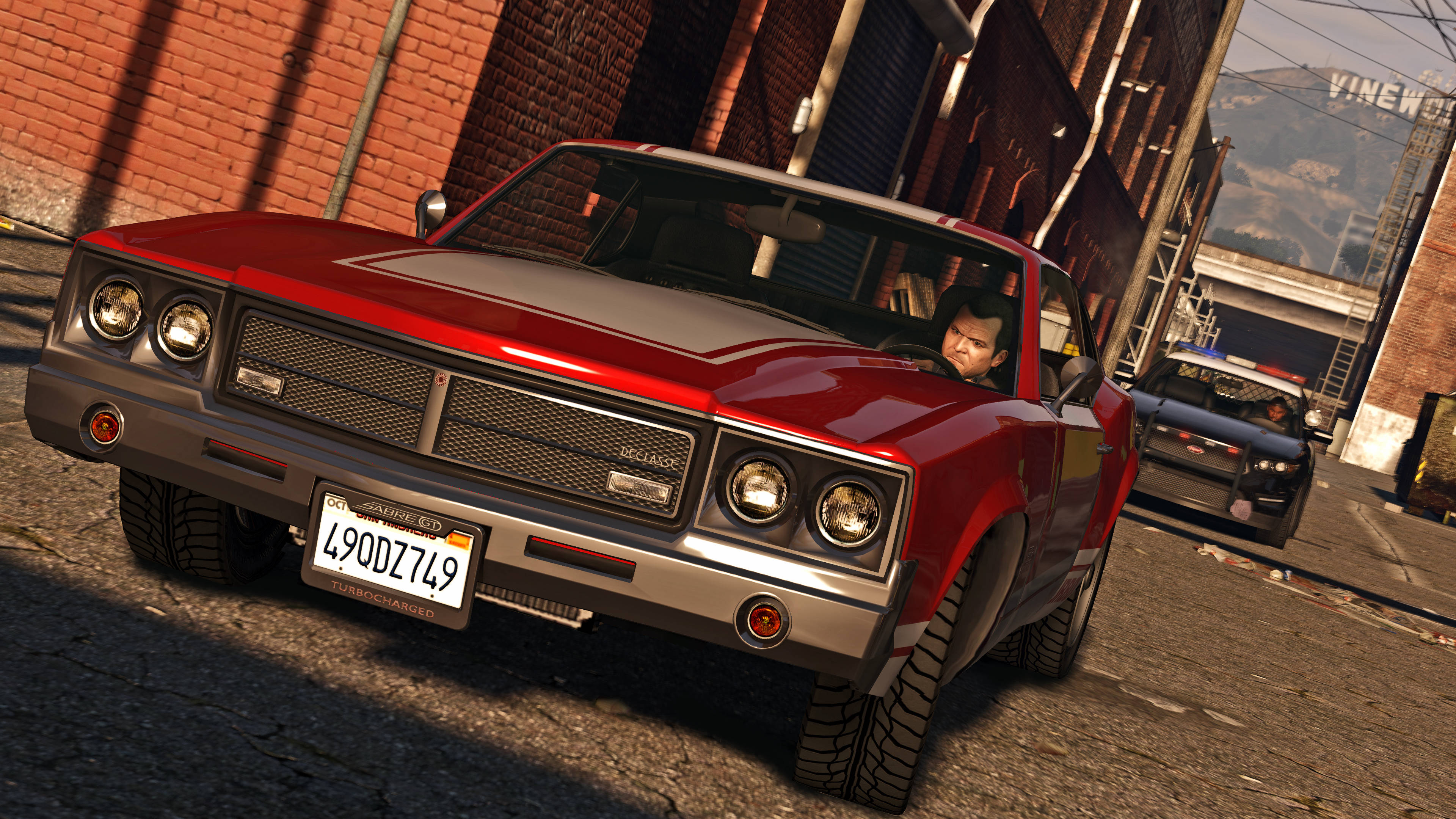 4K gta 5 桌布 壁紙 — 一輛帶有鍍鉻細節的紅色經典肌肉車停放在陽光普照的城市街道上，街道上有磚砌建築，車內有司機。