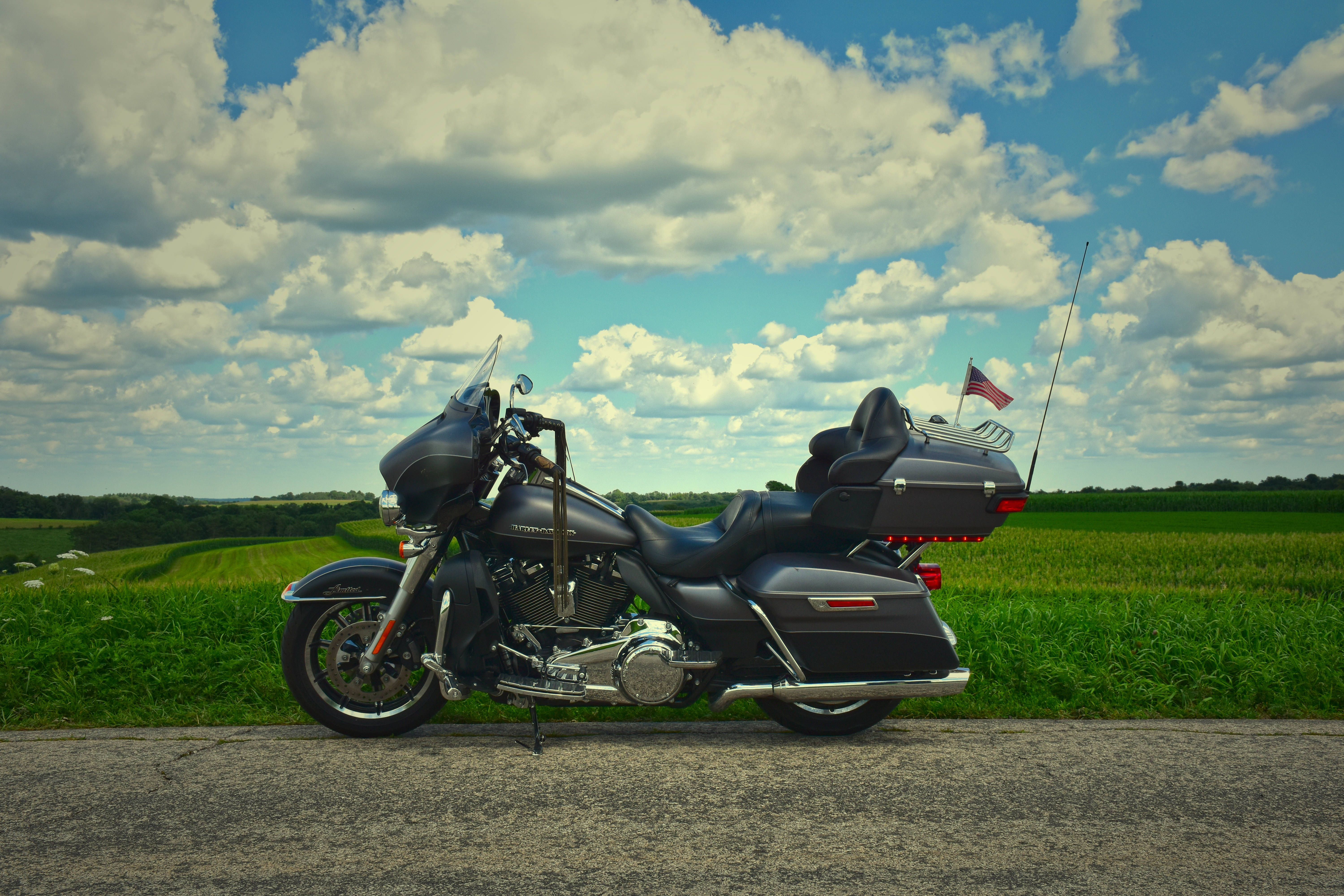 Harley 桌布 壁紙 — 一輛帶有鍍鉻裝飾和美國國旗的黑色旅行摩托車停在鄉村道路上，天空湛藍，白雲朵朵。