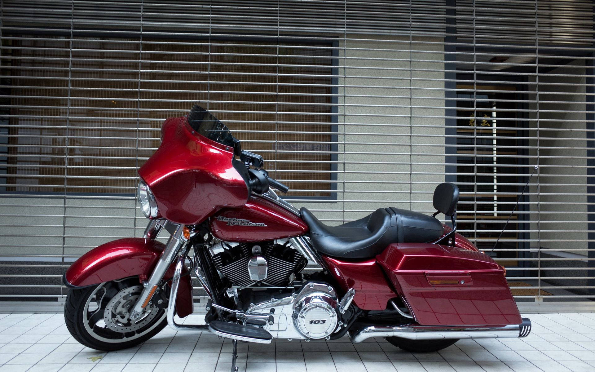 Harley 桌布 壁紙 — 一輛帶有鍍鉻裝飾的亮紅色旅行機車，停放在鋪有瓷磚的地板上，前面是一座帶有水平百葉窗的建築物。