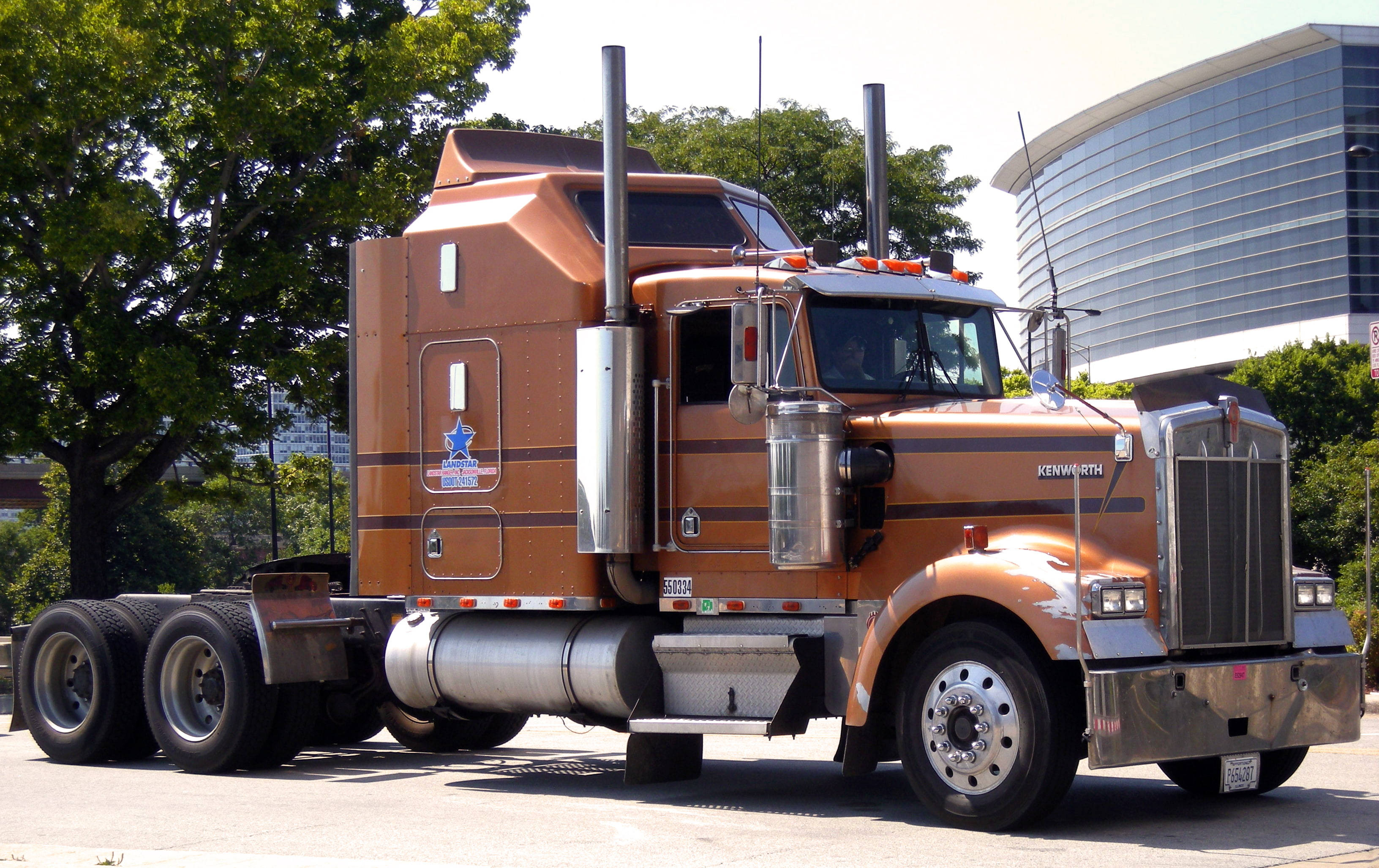 Kenworth W900 桌布 壁紙 — 一輛帶有鍍鉻裝飾的大型橙色Kenworth半掛卡車停在城市街道上，旁邊有綠樹和現代玻璃建築。