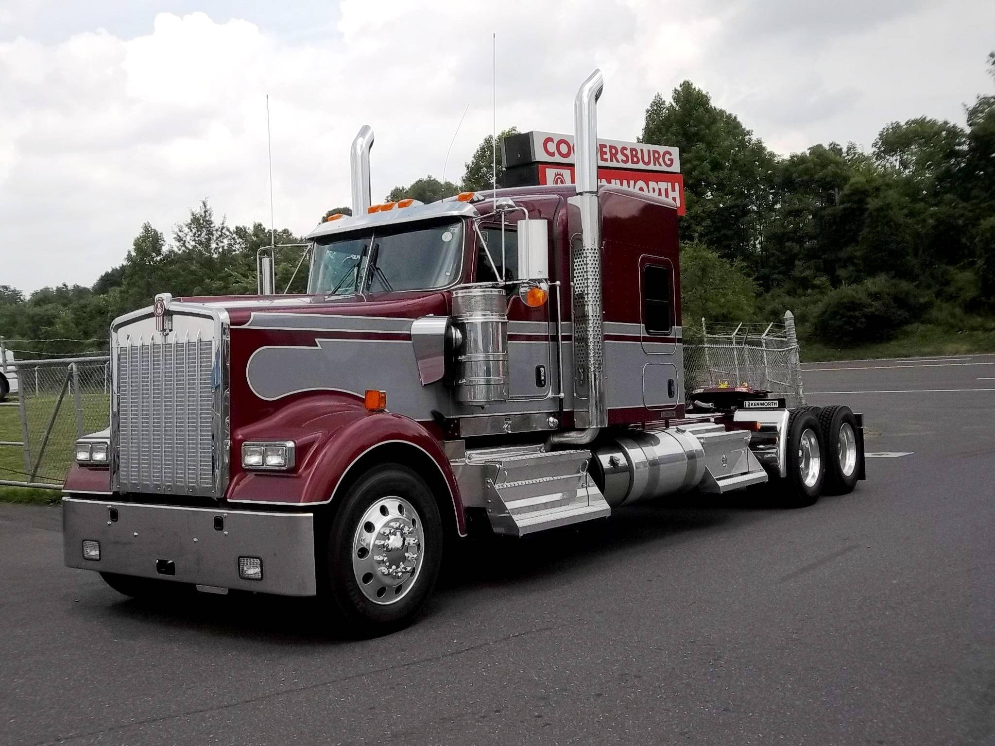 Kenworth W900 桌布 壁紙 — 一輛帶有鍍鉻裝飾的栗色和銀色 Kenworth 半掛卡車停在柏油路上，靠近鐵絲網圍欄和綠樹，天空多雲。