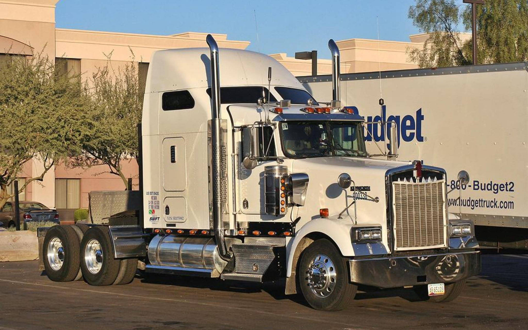 Kenworth W900 桌布 壁紙 — 一輛帶有鍍鉻裝飾的純白色Kenworth半掛卡車停放在陽光普照的停車場，旁邊有一輛Budget拖車。