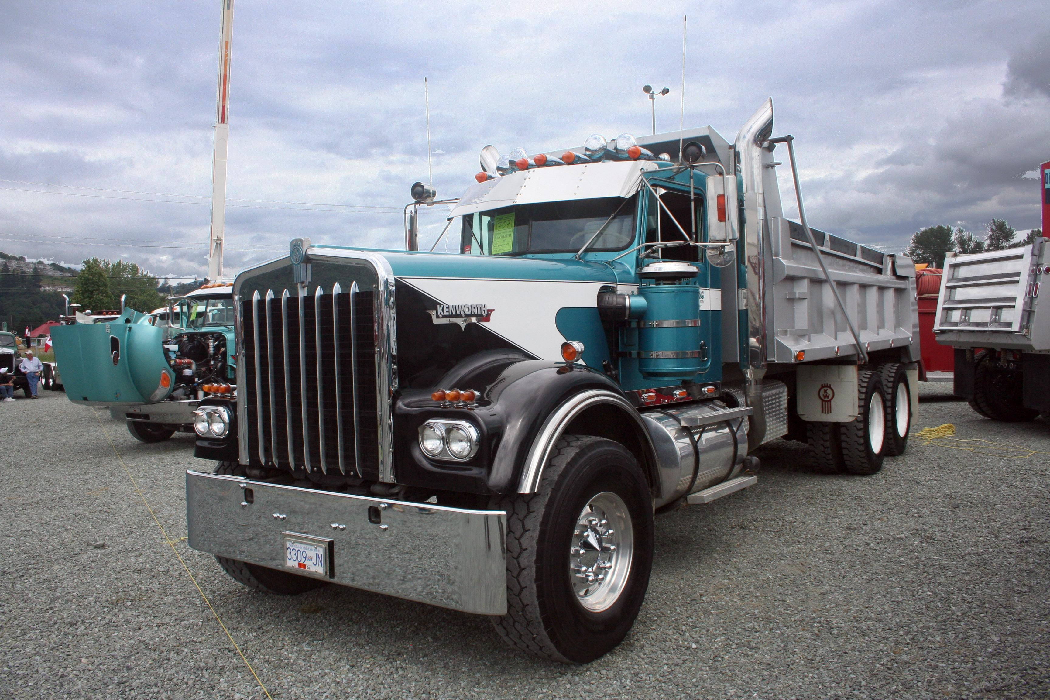 Kenworth W900 桌布 壁紙 — 一輛帶有鍍鉻裝飾的藍綠色和白色Kenworth自卸車在陰天下的礫石上顯眼地停放。