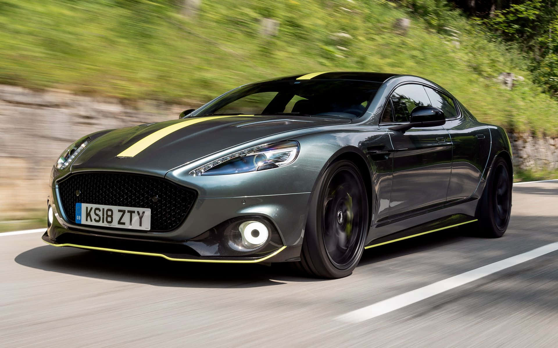 Aston Martin Rapide S 桌布 壁紙 — 一輛帶有黃色點綴的時尚灰色阿斯頓馬丁在蜿蜒的道路上穿過鬱鬱蔥蔥的綠色山丘飛馳。