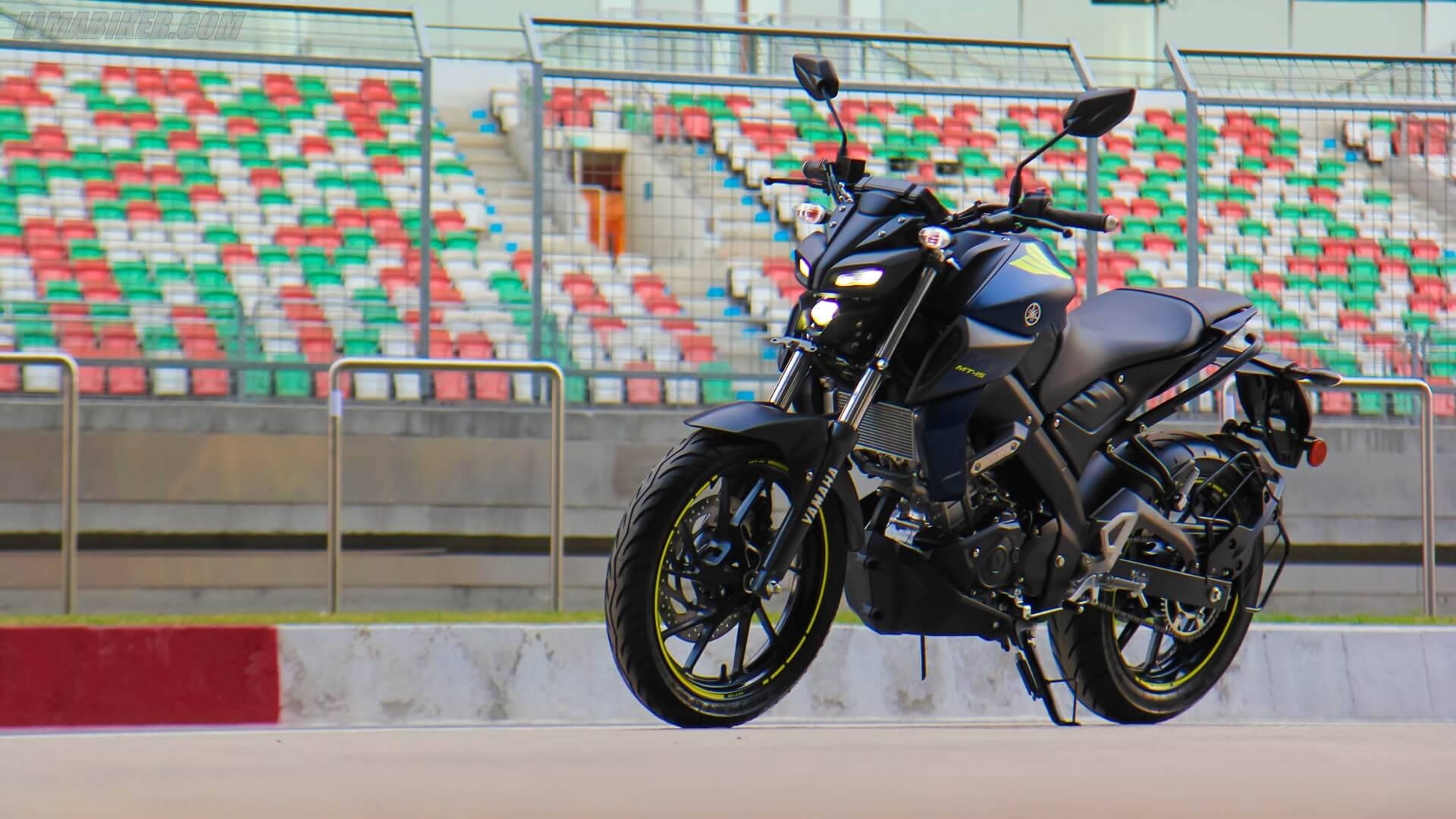 yamaha mt 15 桌布 壁紙 — 一輛帶有黃色點綴的時尚黑色運動摩托車，在賽道上停放在充滿活力的紅色、綠色和白色體育場座位旁。