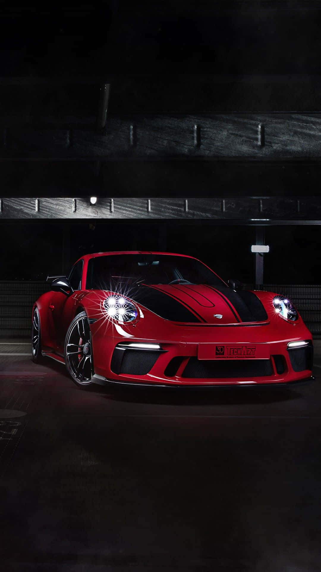 Porsche iPhone 桌布 壁紙 — 一輛帶有黑色條紋的光澤紅色跑車在黑暗的工業車庫中於聚光燈下閃閃發光。