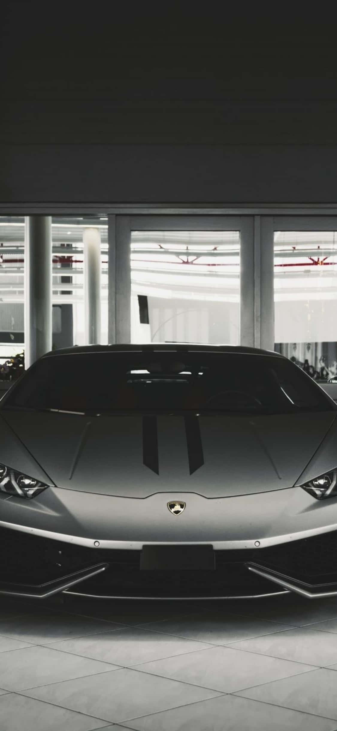iPhone X Lamborghini 桌布 壁紙 — 一輛帶有黑色條紋的時尚銀色藍寶堅尼跑車，位於現代簡約的展廳中央，地板舖有瓷磚，並設有大窗戶。
