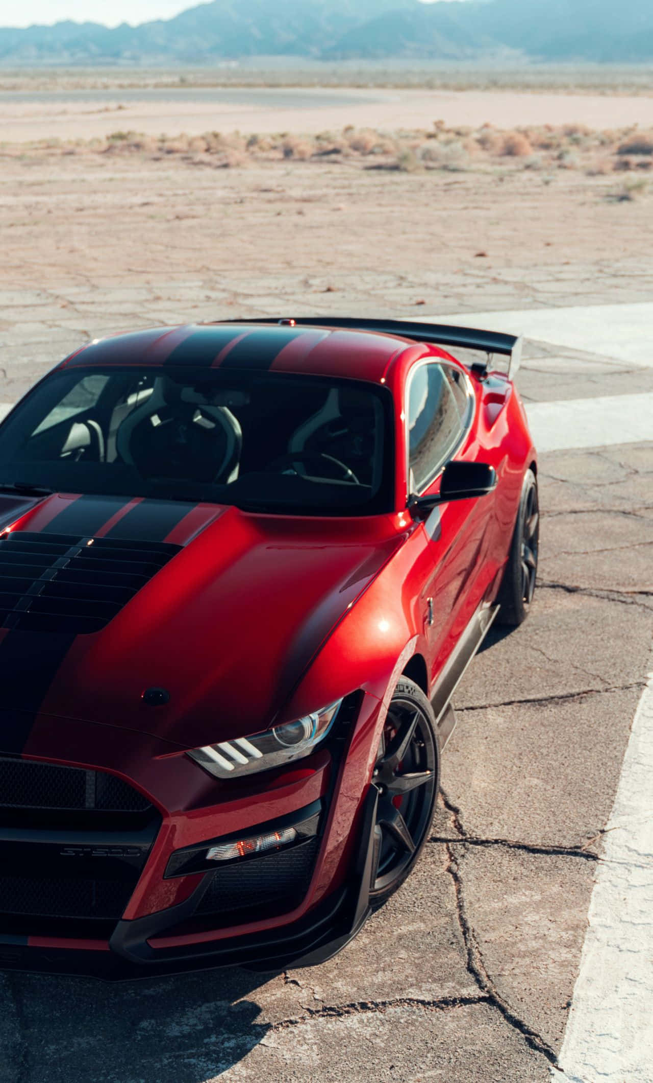 Mustang iPhone 桌布 壁紙 — 一輛帶有黑色賽車條紋的亮紅色跑車停在荒涼沙漠景觀中一條破裂的瀝青賽道上。
