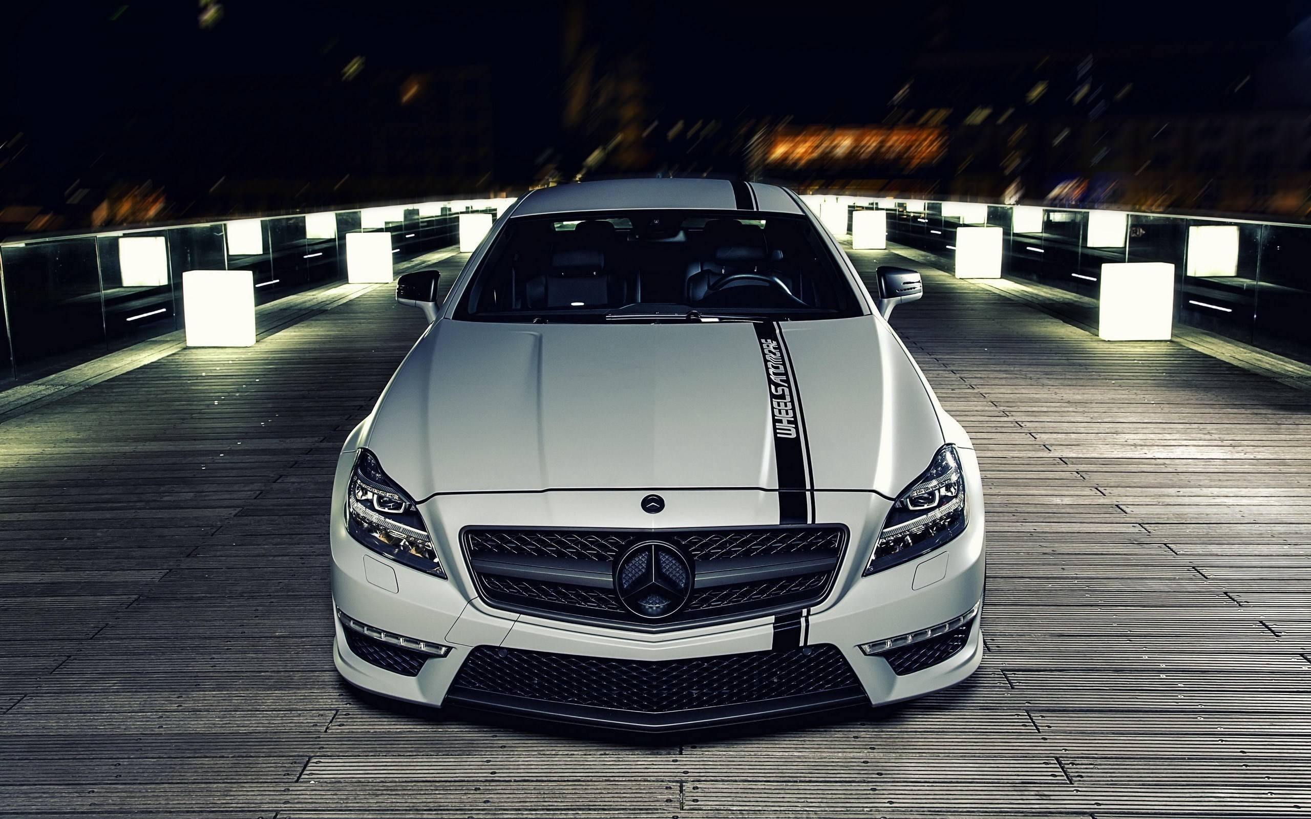 Mercedes Benz Amg 桌布 壁紙 — 一輛帶有黑色賽車條紋的時尚白色賓士跑車，夜間位於木橋中央，由發光的白色立方體和模糊的城市燈光照亮。