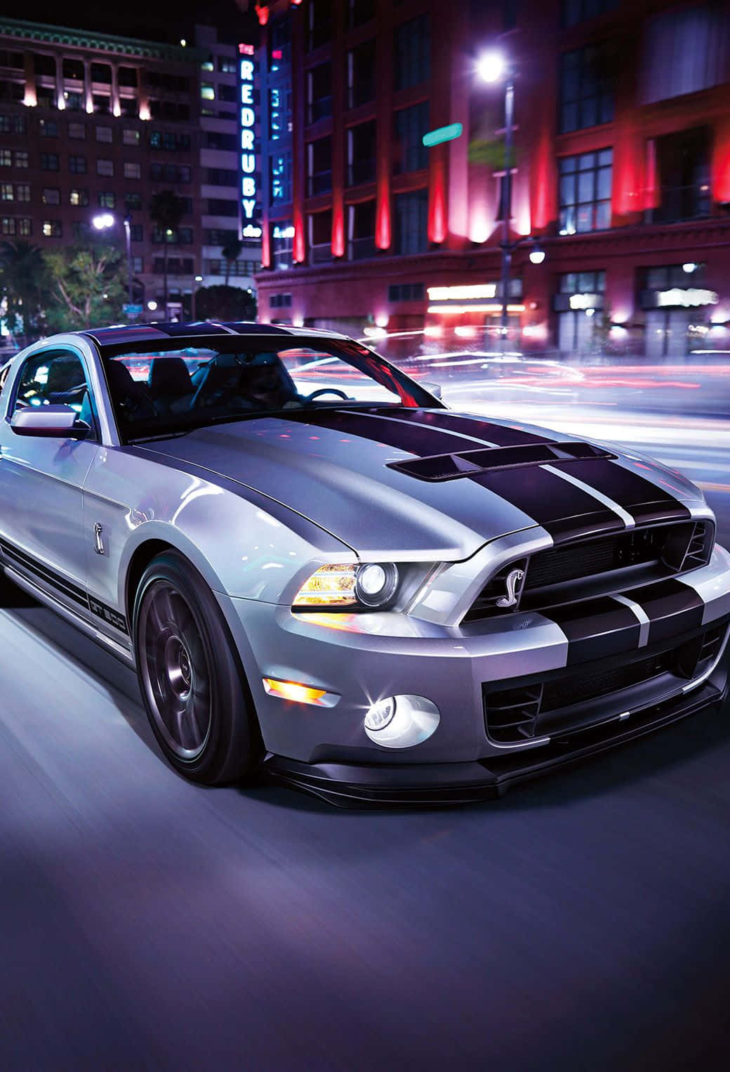 Ford Mustang 桌布 壁紙 — 一輛帶有黑色賽車條紋的銀色福特野馬在夜間穿過霓虹燈照亮的大街，留下模糊的運動軌跡。