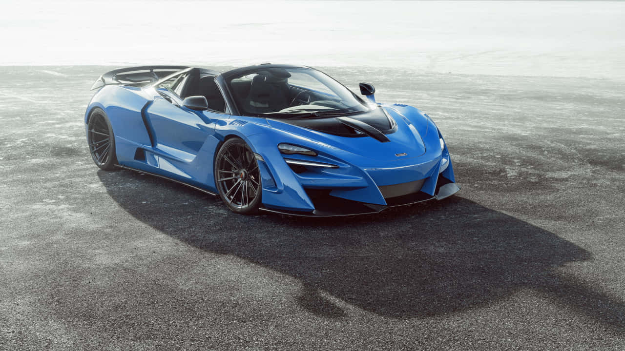 汽車 桌布 壁紙 — 一輛帶有黑色點綴的時尚藍色McLaren超級跑車，在明亮的日光下停放在灰色瀝青路面上。