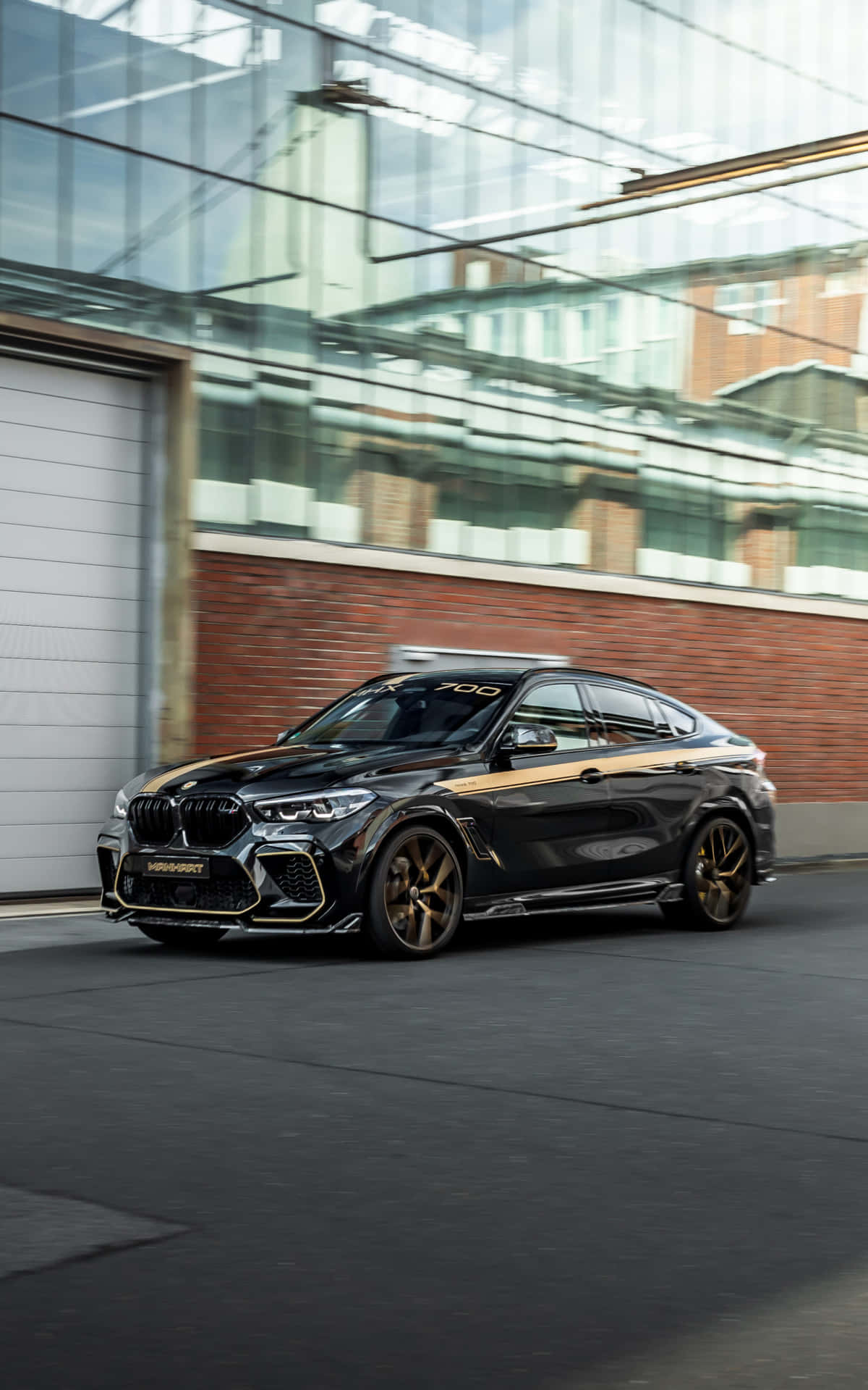 Bmw X6 M 桌布 壁紙 — 一輛帶金色裝飾的時尚黑色BMW X6 M700在城市的街道上飛馳而過，經過一座現代化的玻璃和磚砌建築。
