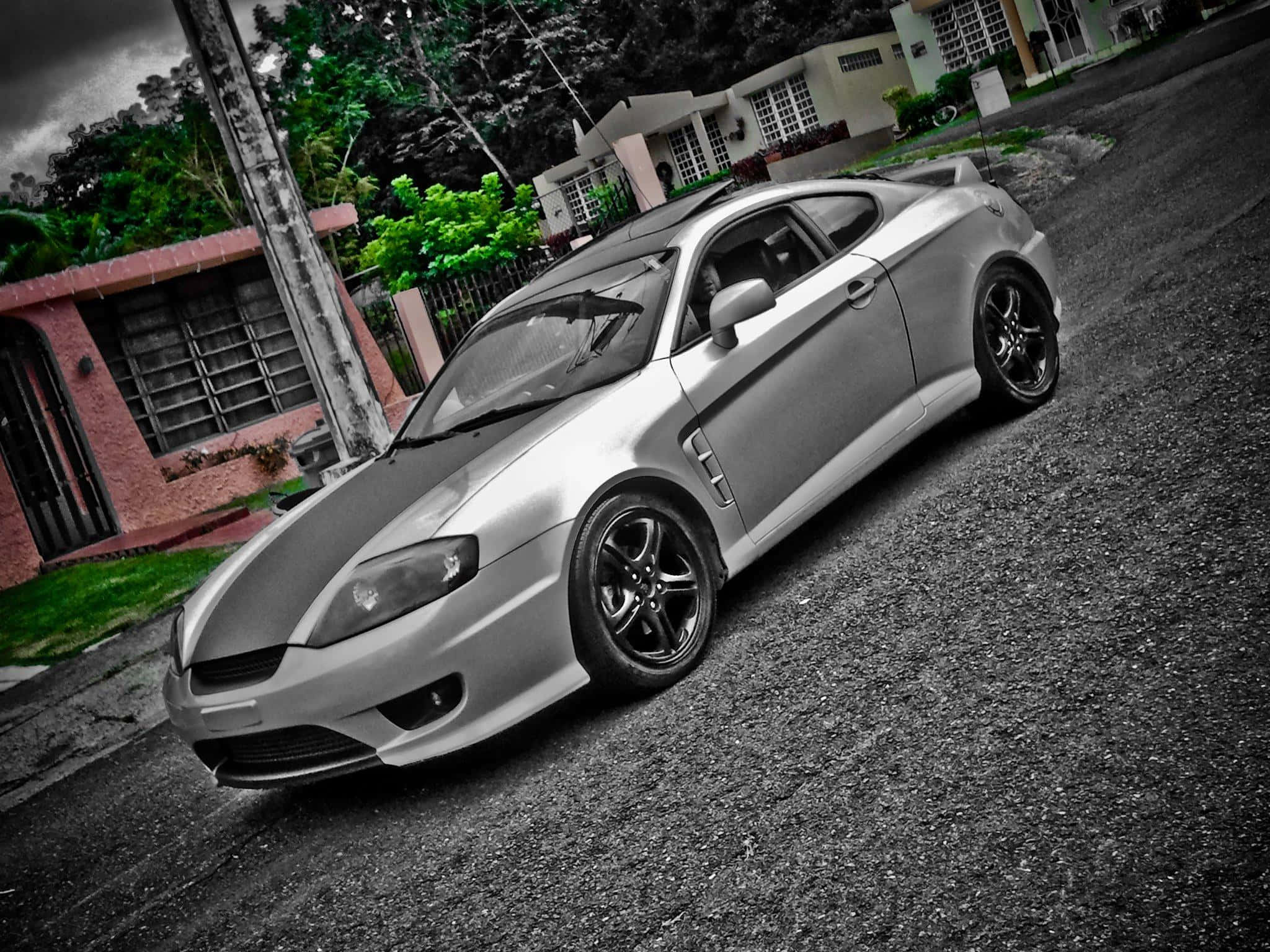 Hyundai Tiburon 桌布 壁紙 — 一輛帶黑色裝飾的銀色跑車停放在碎石車道上，旁邊是一棟粉紅色的房子，天空多雲。