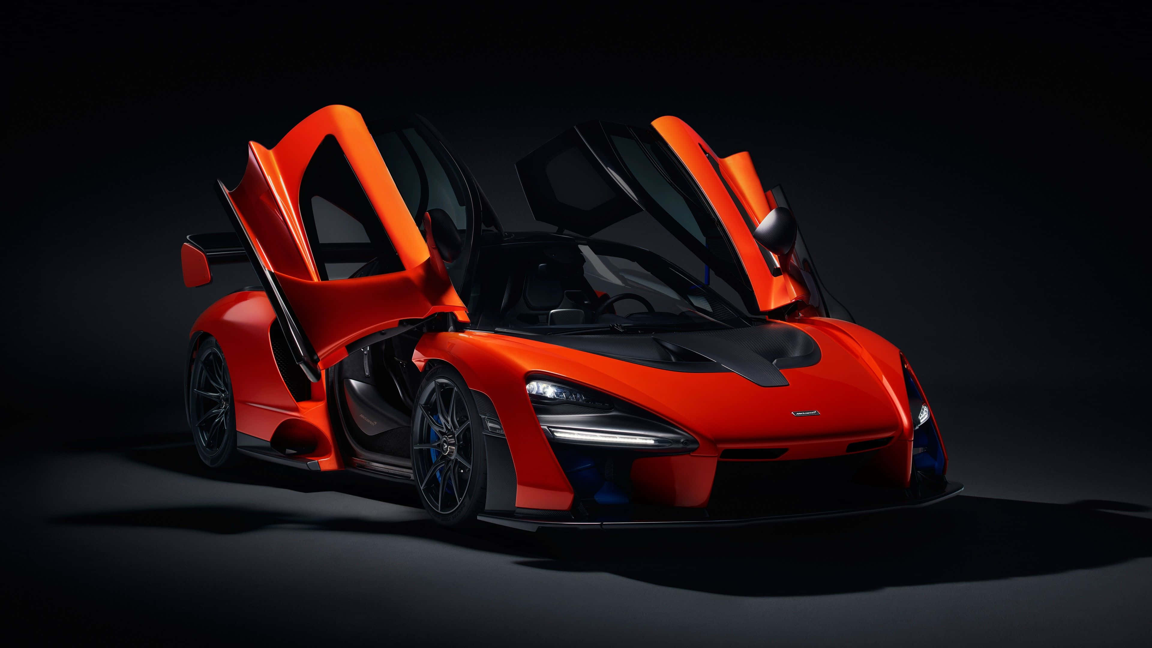 Cj So Cool 桌布 壁紙 — 一輛引人注目的橙色McLaren Senna超級跑車，鷗翼車門打開，在黑色背景下被戲劇性的工作室燈光照亮。