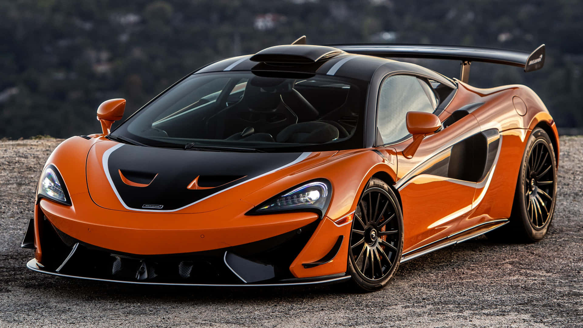 Mclaren 620r 桌布 壁紙 — 一輛引人注目的橙色和黑色McLaren超級跑車，帶有大型後擾流板，停在碎石山坡上。