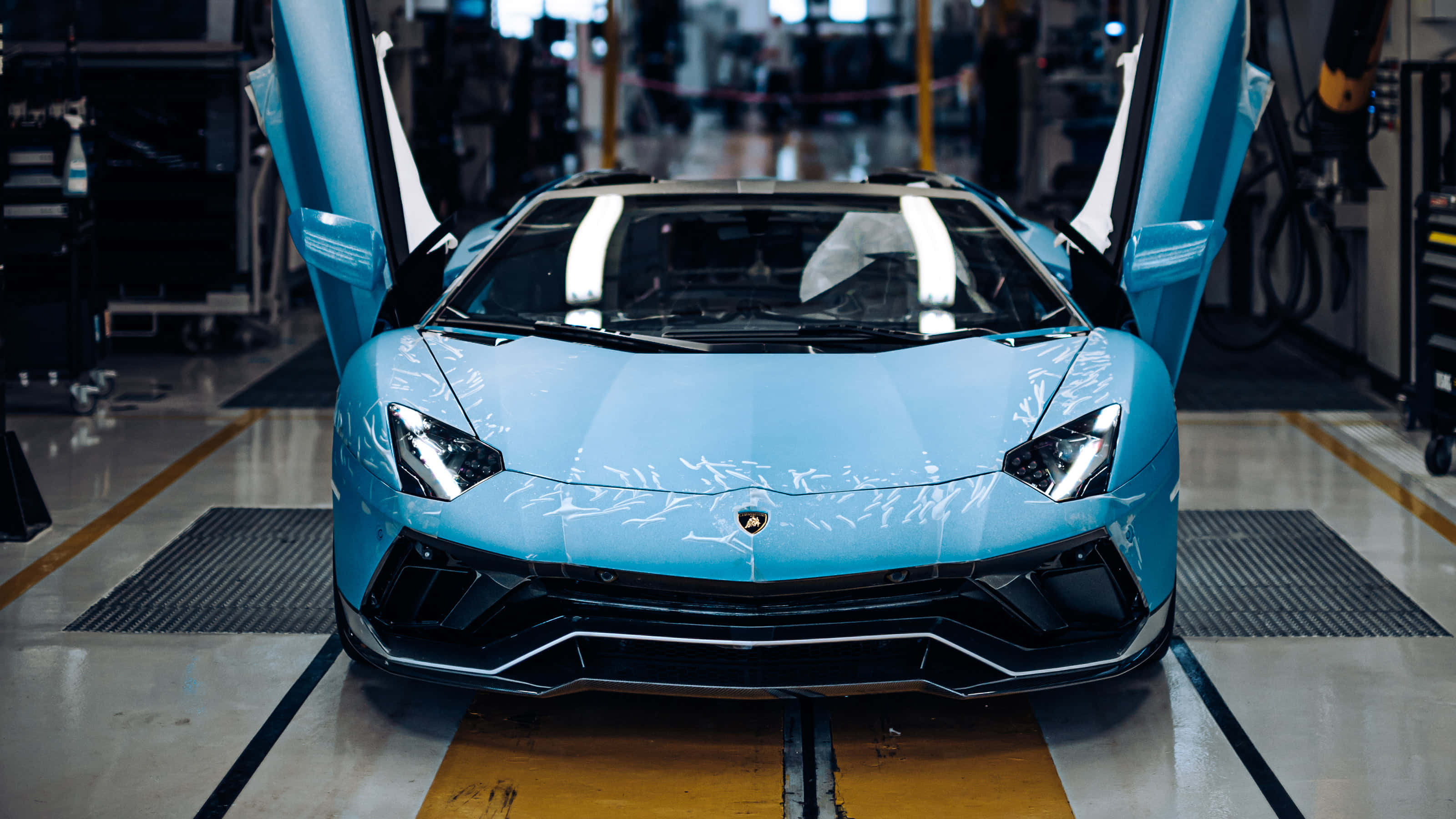 Lamborghini Aventador 桌布 壁紙 — 一輛引人注目的淺藍色藍寶堅尼，剪刀門打開，在明亮的工業燈光下自豪地站在工廠地板上。