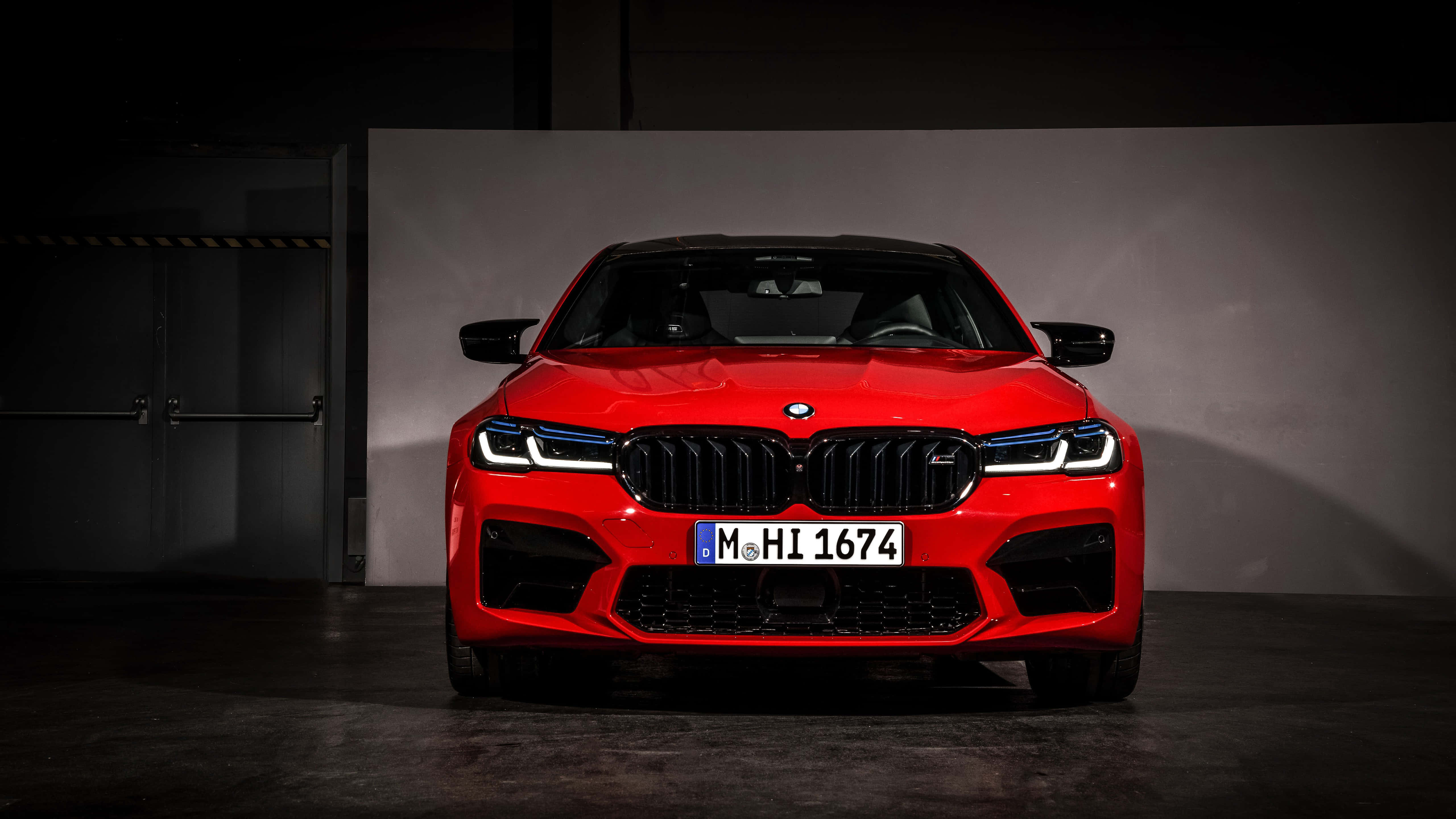 桌布 壁紙 — 一輛引人注目的紅色BMW M5跑車在黑暗的工業車庫中從正面戲劇性地被照亮。