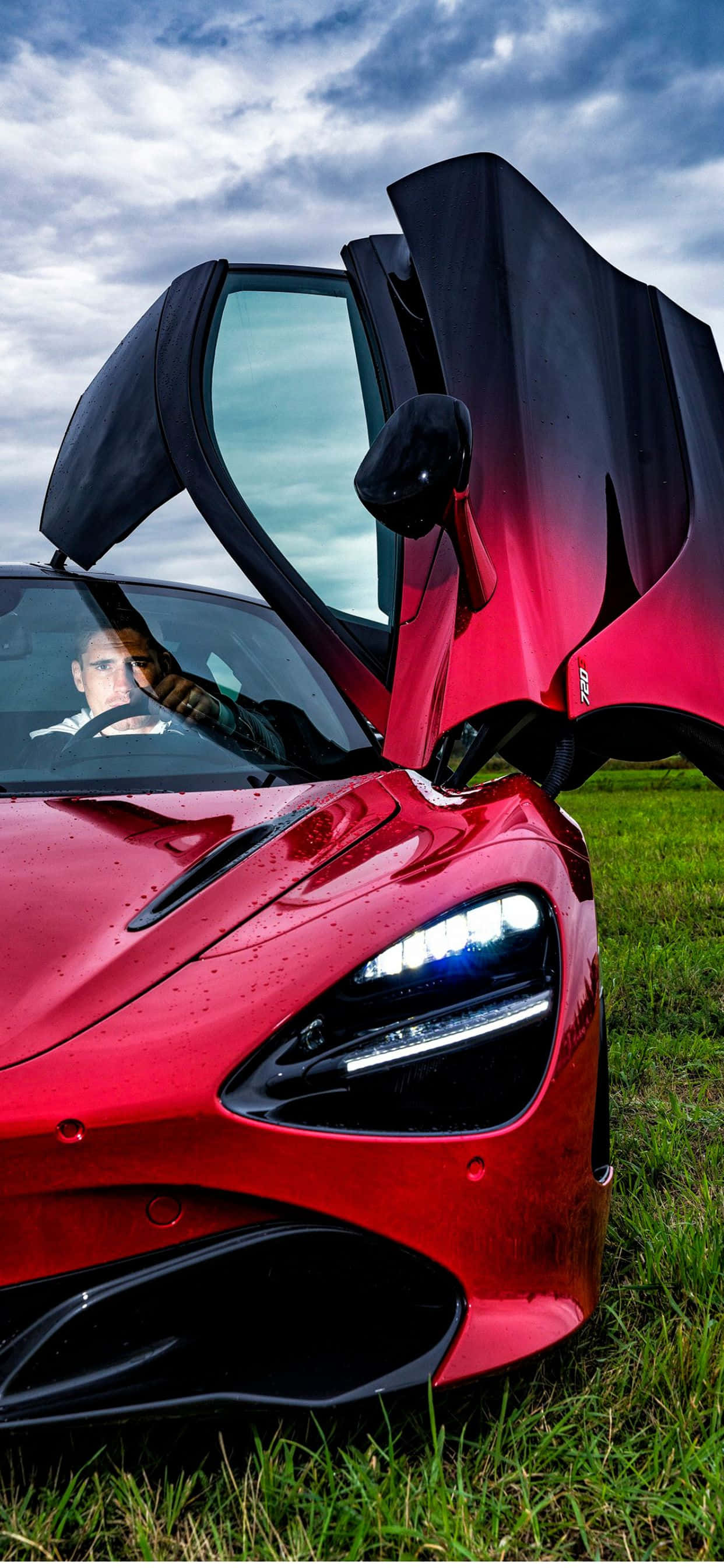 黑色 桌布 壁紙 — 一輛引人注目的紅色McLaren跑車，鷗翼門打開，停在草地上，天空多雲，車內有駕駛。