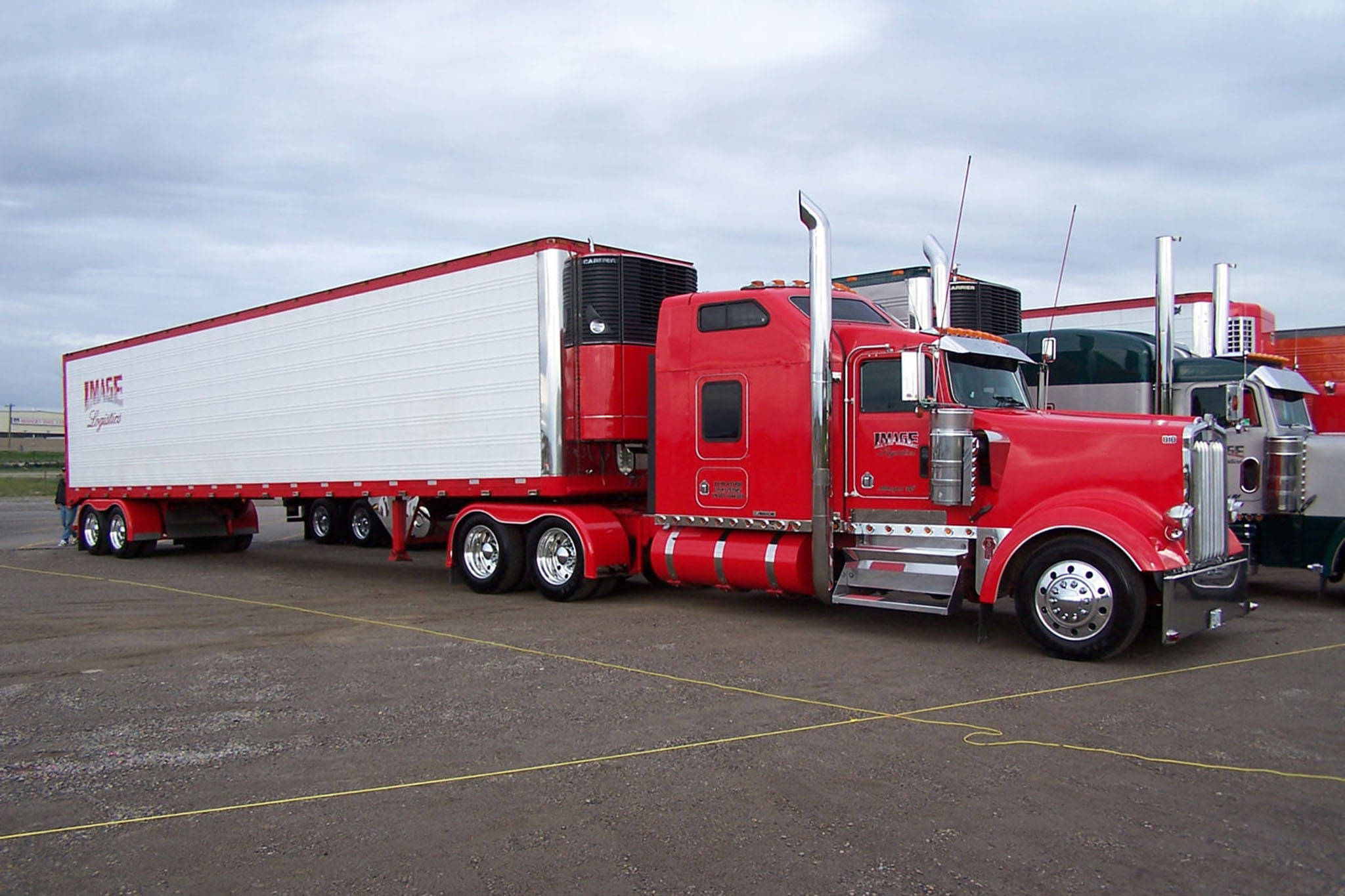 Kenworth W900 桌布 壁紙 — 一輛引人注目的紅色半掛卡車帶白色拖車停在灰色瀝青場地上，天空多雲。
