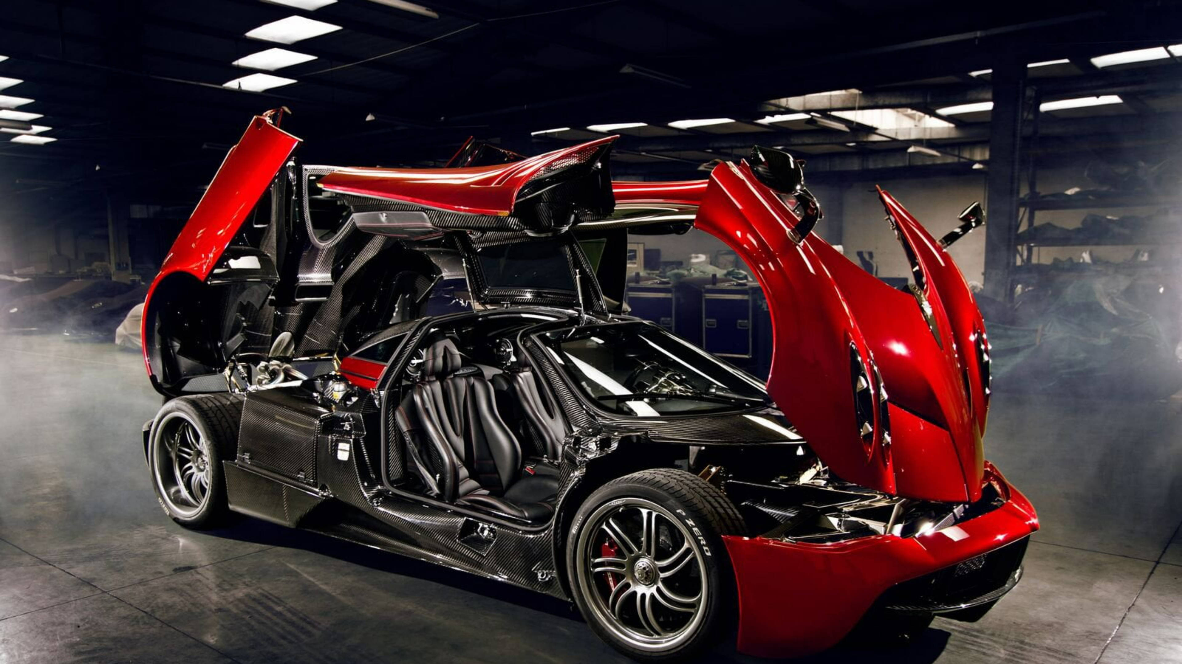 Pagani 桌布 壁紙 — 一輛引人注目的紅色和黑色帕加尼Huayra，剪刀門打開，停在煙霧瀰漫的工業車庫中，上方有明亮的燈光。