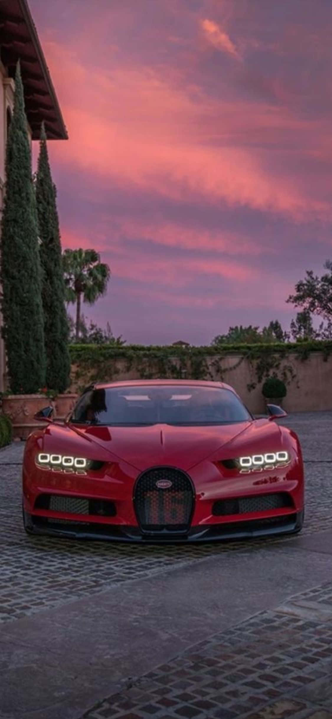 iPhone Xs Bugatti 桌布 壁紙 — 一輛引人注目的紅色布加迪Chiron停在鵝卵石車道上，背景是戲劇性的粉紅和紫色日落天空。