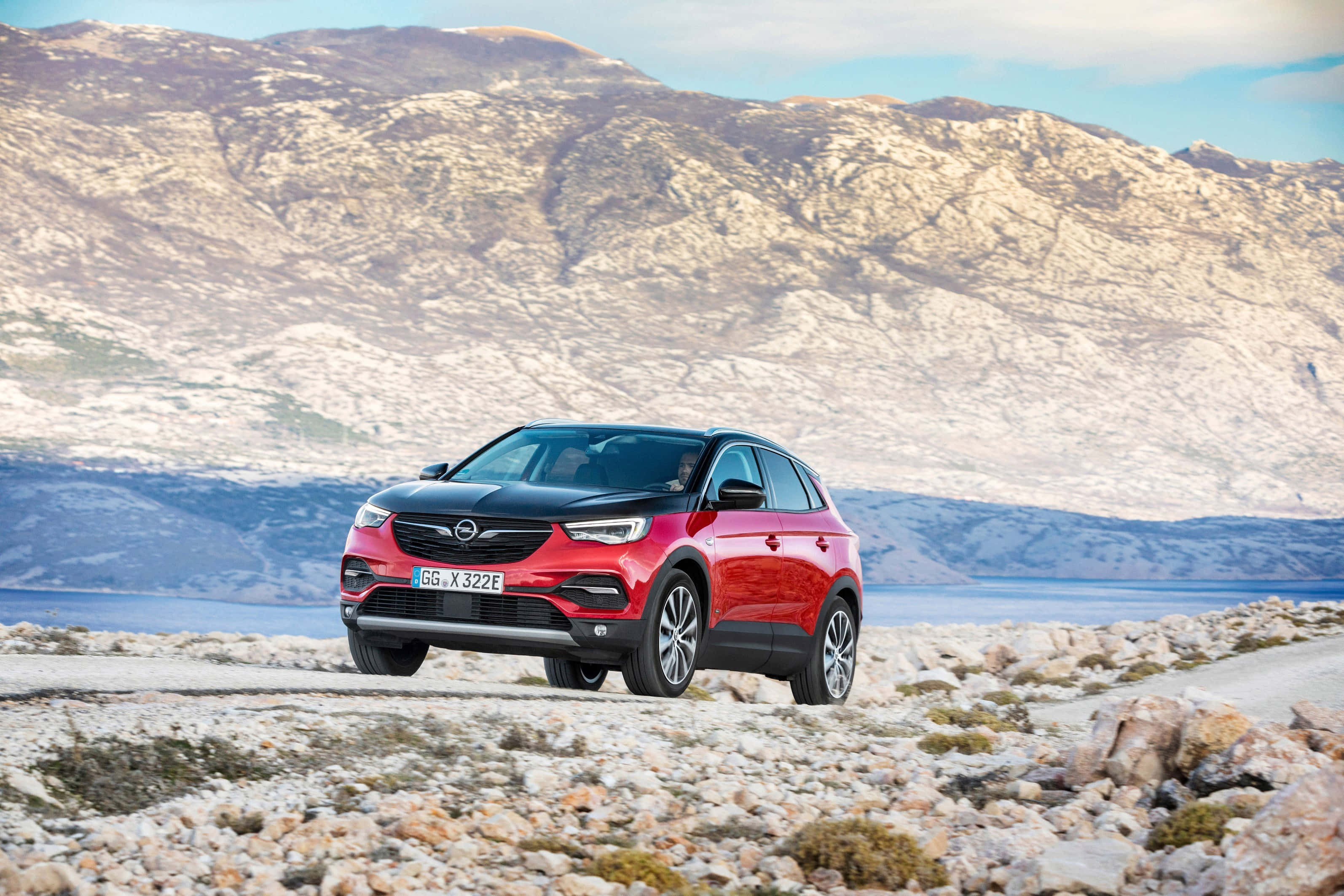Opel Grandland X 桌布 壁紙 — 一輛引人注目的紅黑SUV停放在岩石海岸道路上，背景是崎嶇的山脈和寧靜的大海。