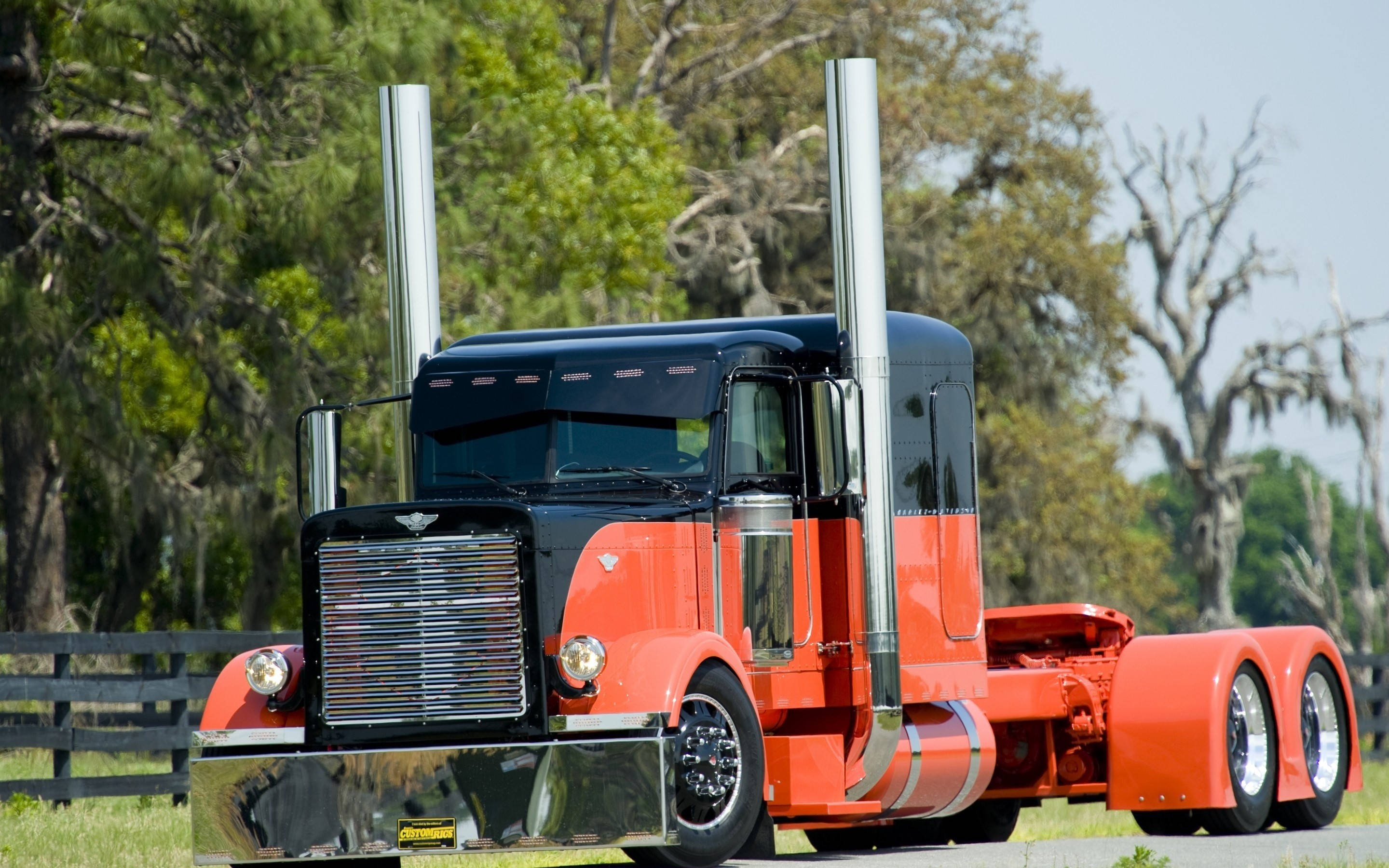 Peterbilt 桌布 壁紙 — 一輛引人注目的紅黑色半掛卡車，帶有鍍鉻排氣管，停在鄉村道路上，旁邊是木柵欄和枝葉茂密的樹木。