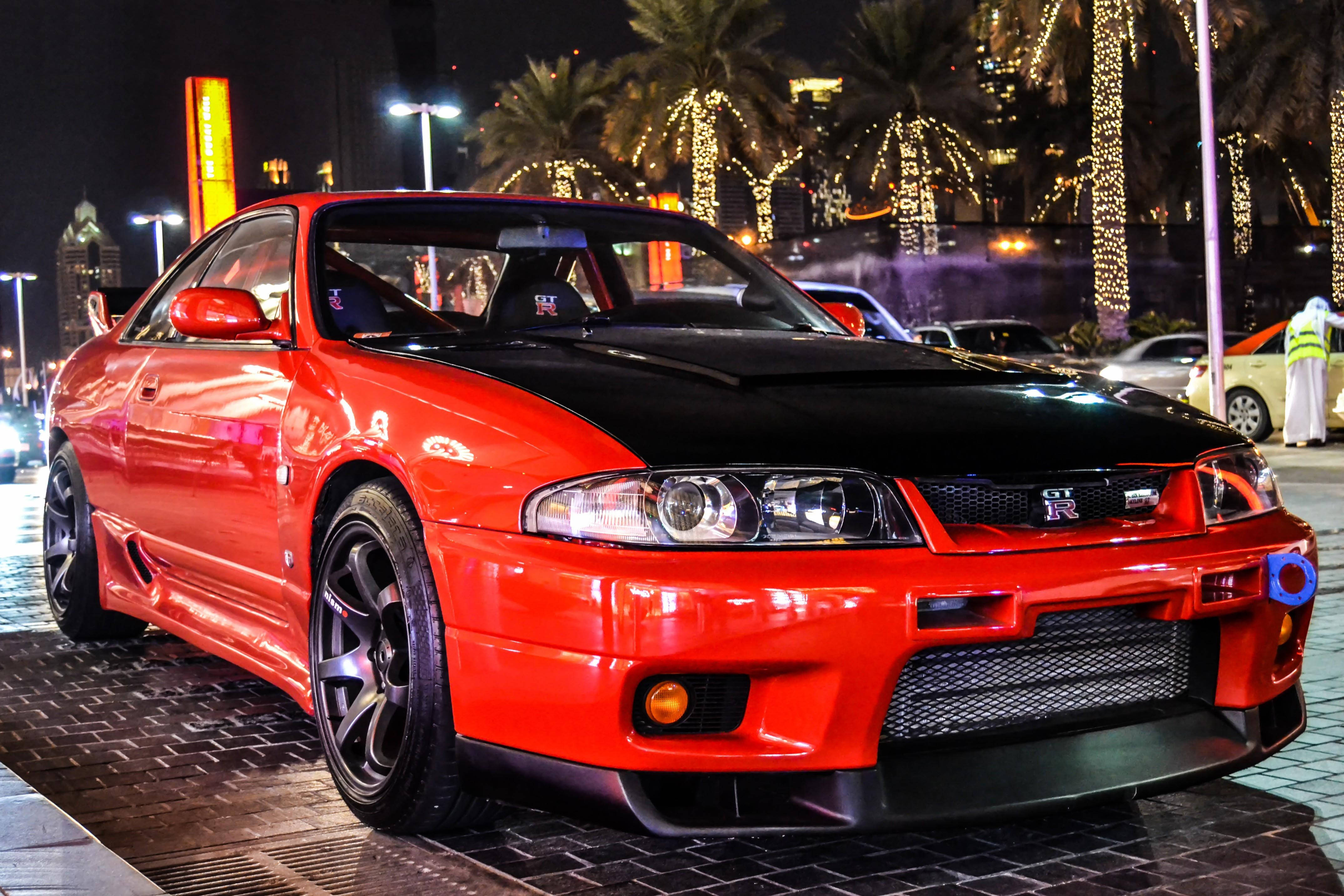 Gtr 桌布 壁紙 — 一輛引人注目的紅黑配色日產GT-R跑車在夜晚的城市燈光下閃閃發光，背景有棕櫚樹和節日燈飾。