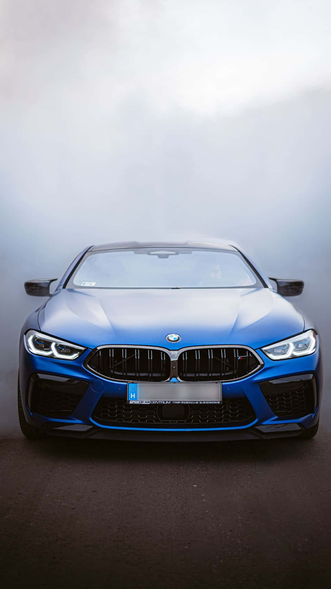 Android Bmw 桌布 壁紙 — 一輛引人注目的藍色BMW M8 Coupe在柔和光線下閃閃發光，其侵略性的前格柵和鋒利的頭燈主導了迷霧、極簡的工作室環境。