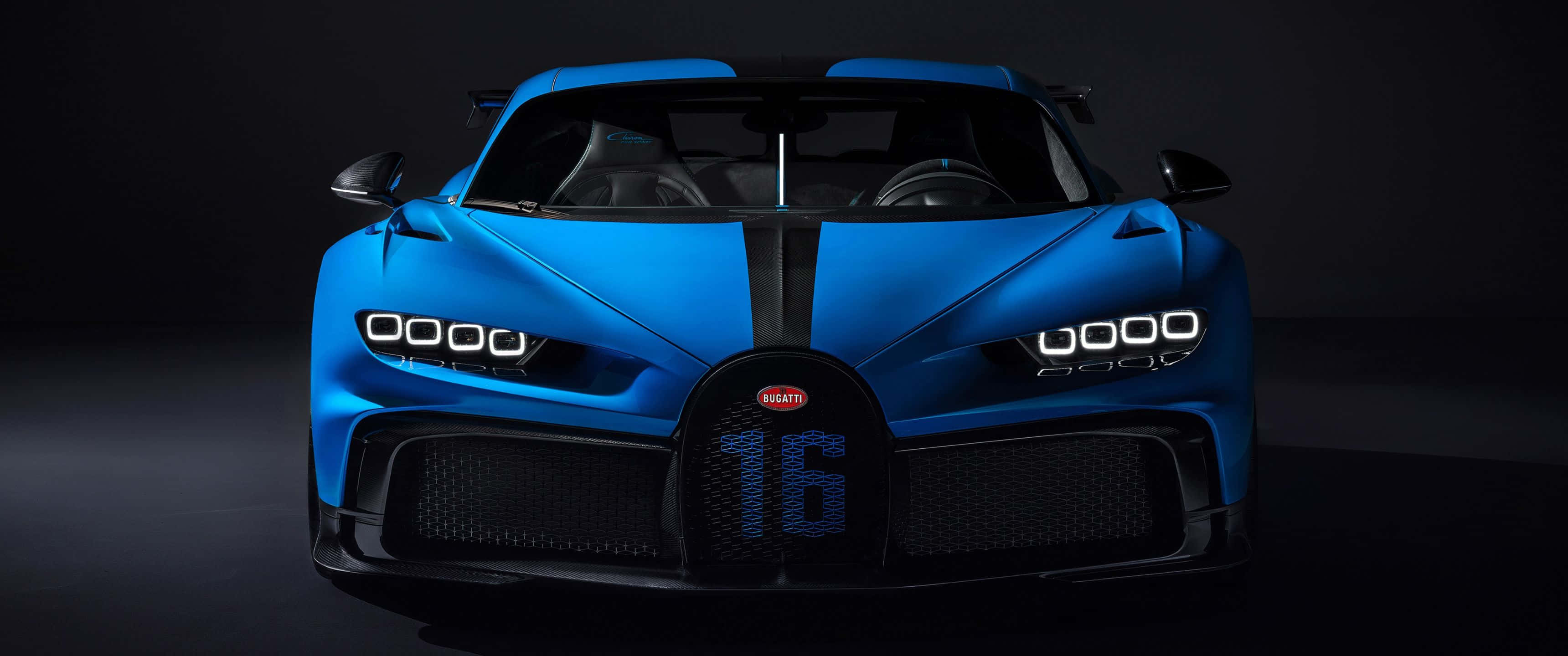 3440x1440p Bugatti 桌布 壁紙 — 一輛引人注目的藍色Bugatti Chiron Super Sport 300+，前格柵上有16號標誌，在黑暗的攝影棚背景下被銳利的車頭燈照亮。
