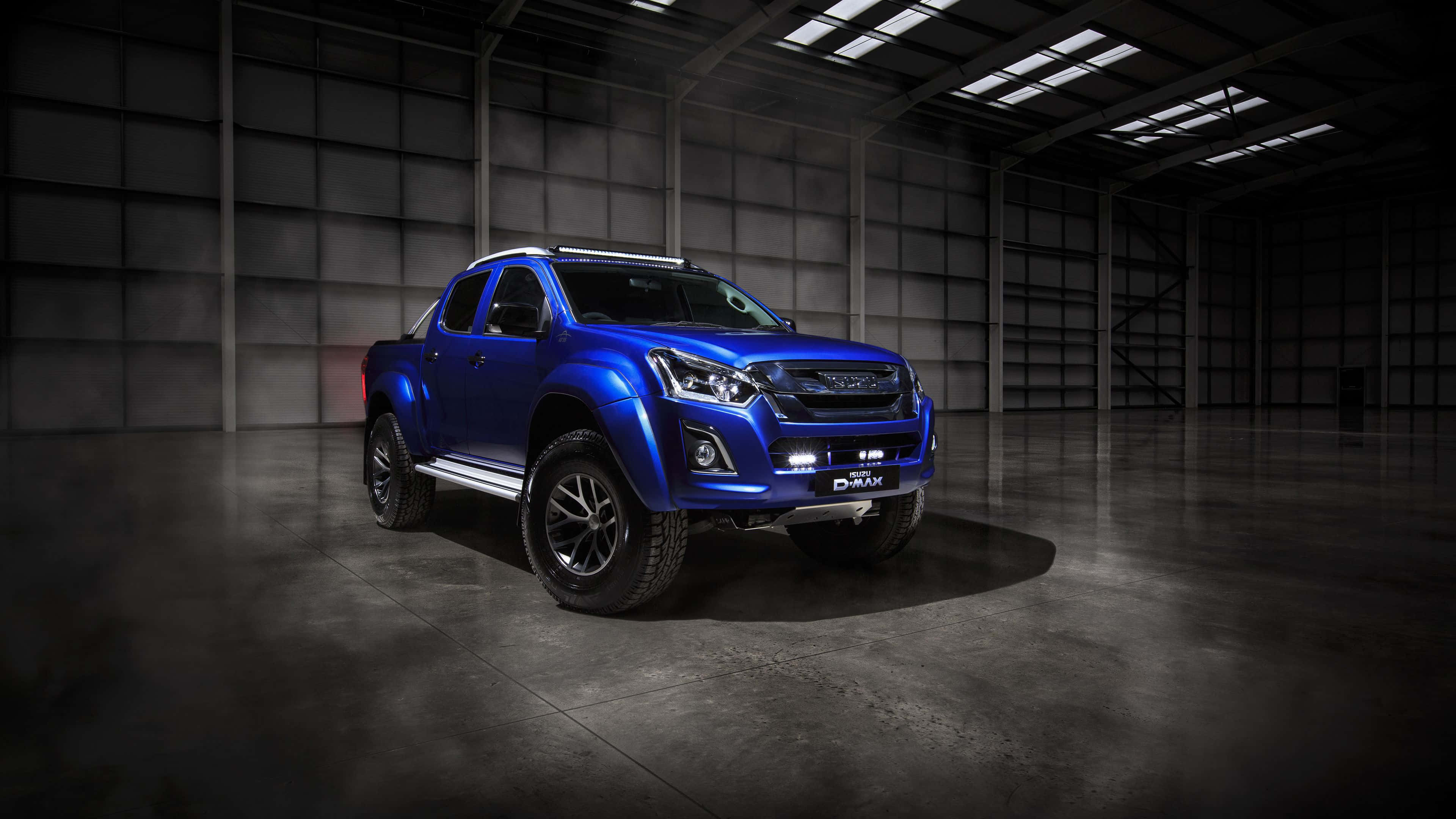 Isuzu 桌布 壁紙 — 一輛引人注目的藍色Isuzu D-Max皮卡車在燈光昏暗的工業倉庫中展示，帶有戲劇性的聚光燈效果。