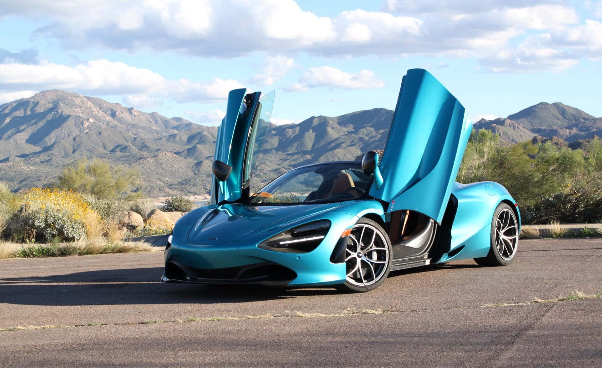 麥拉倫 桌布 壁紙 — 一輛引人注目的青綠色McLaren跑車，鷗翼車門打開，停在沙漠公路上，背景是崎嶇的山脈和部分多雲的天空。