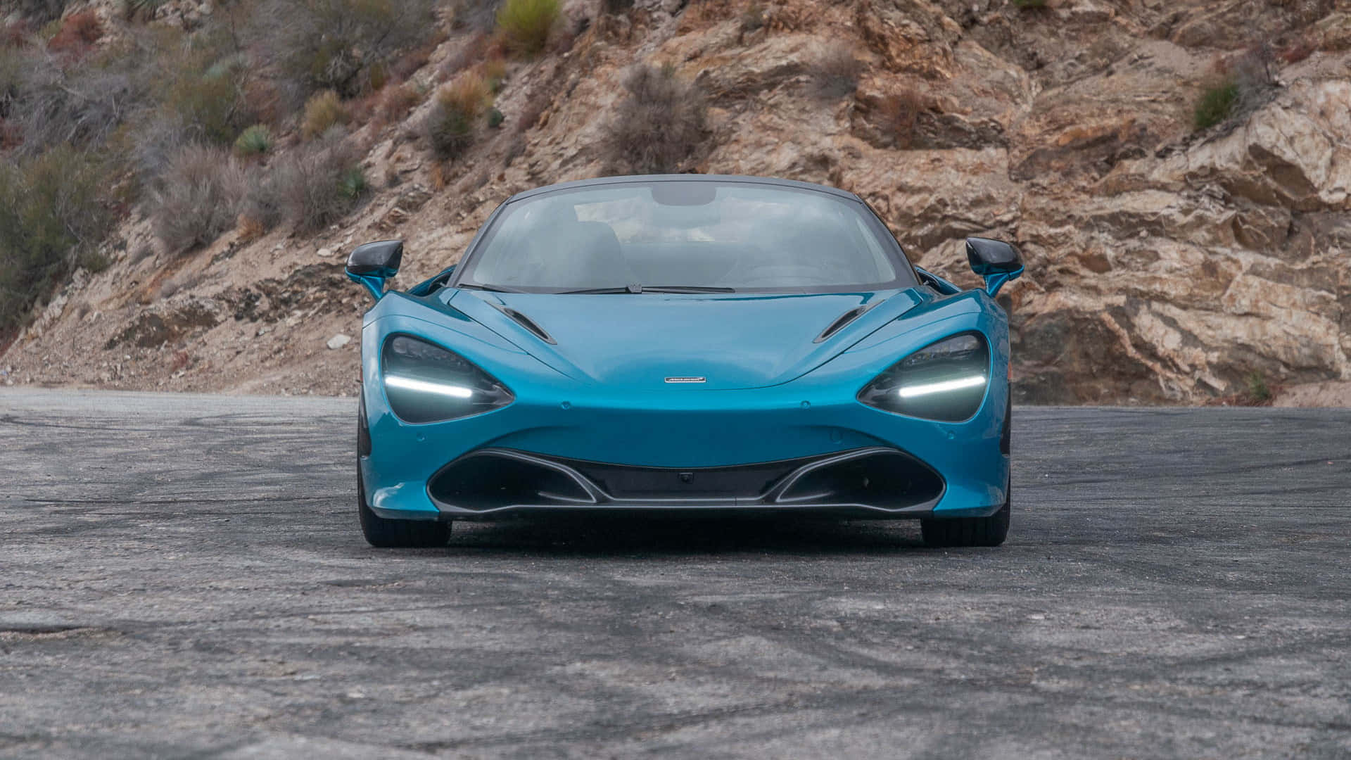 Blue Car 桌布 壁紙 — 一輛引人注目的青綠色McLaren 720S跑車在崎嶇的山路上向前行駛，背景是岩石峭壁和乾燥的灌木叢。
