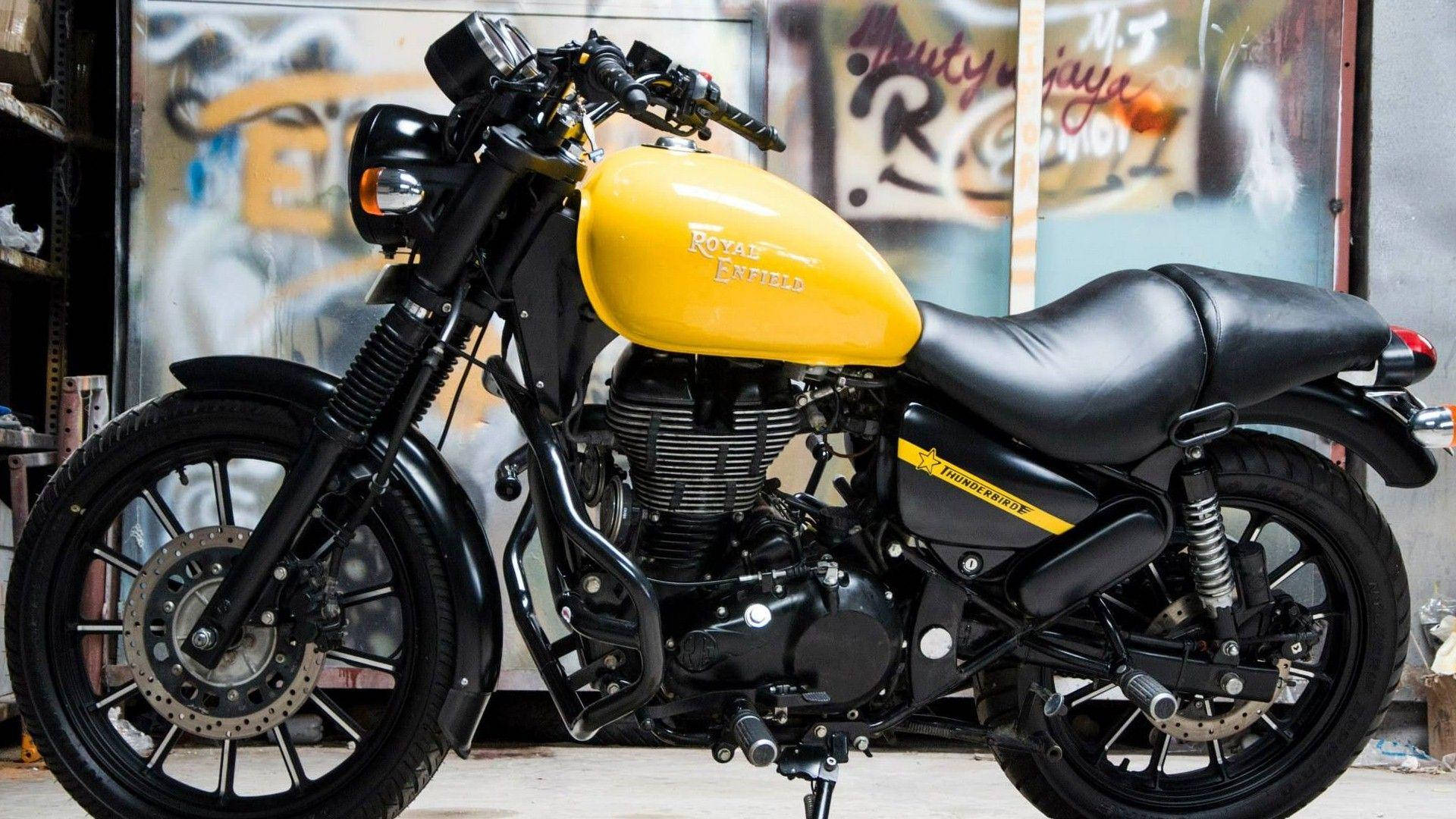 Royal 桌布 壁紙 — 一輛引人注目的黃黑配色 Royal Enfield Thunderbird 摩托車，停放在一個佈滿塗鴉的車庫中，配以工業照明。