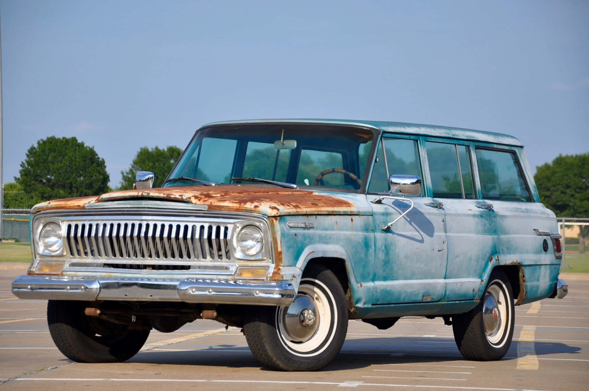 Jeep Wagoneer 桌布 壁紙 — 一輛引擎蓋生鏽的復古藍色旅行車，停在陽光明媚、綠樹環繞的空地上。