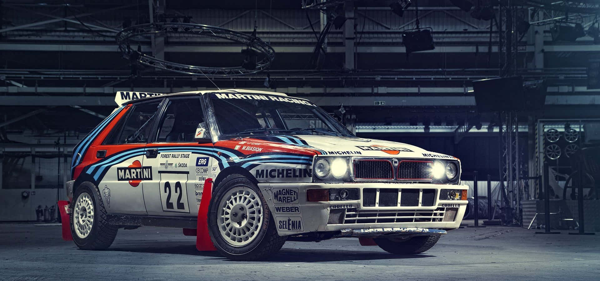 Lancia 桌布 壁紙 — 一輛復古 Martini Racing Lancia Delta S4 拉力賽車，編號 22，停放在光線昏暗的工業車庫內，車庫有裸露的橫樑和頭頂燈光。