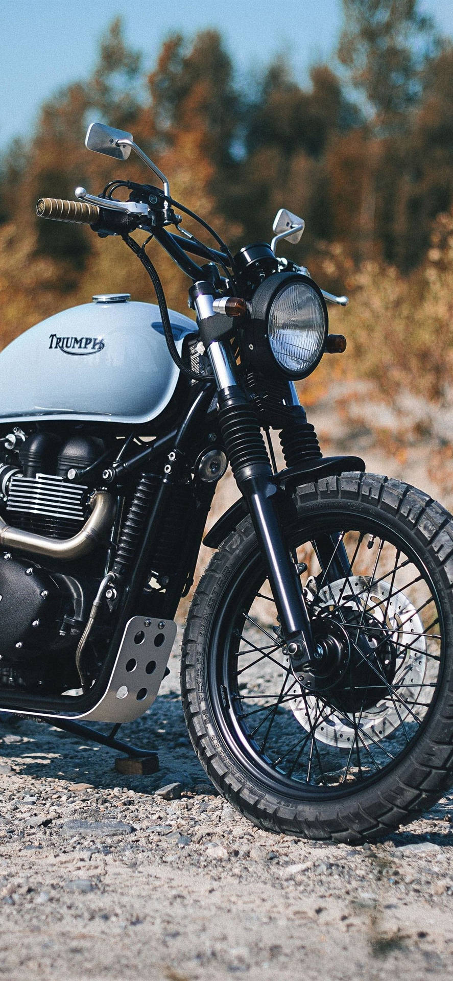 腳踏車 桌布 壁紙 — 一輛復古Triumph摩托車，銀色油箱和黑色車架，站在碎石上，背景是晴朗的藍天和秋天的樹木。
