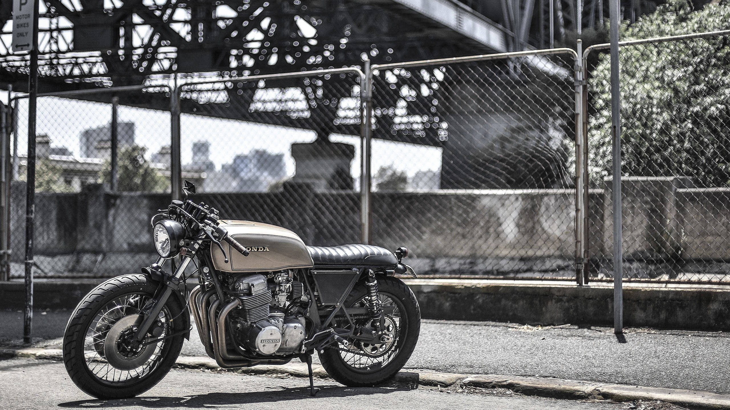 Bobber 桌布 壁紙 — 一輛復古咖啡賽車摩托車停放在鋼橋下，背景有鐵絲網圍欄和城市天際線。
