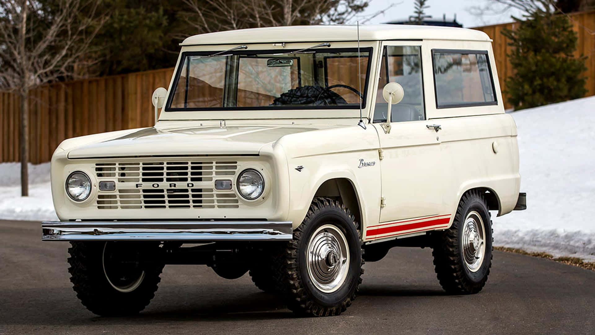 福特野馬越野車 桌布 壁紙 — 一輛復古奶油色福特Bronco，帶有紅色條紋，停在雪地路邊，附近有光禿禿的樹木。
