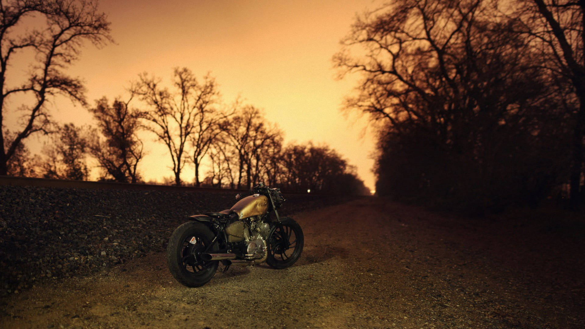 Bobber 桌布 壁紙 — 一輛復古摩托車停在碎石路上，夕陽西下，在溫暖的橙色天空下，被光禿禿的樹木環繞。