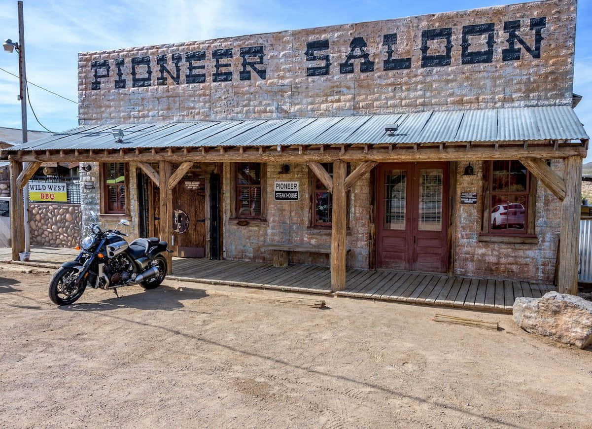 Old Western 桌布 壁紙 — 一輛復古摩托車停泊在樸實的石頭沙龍前，上方有 'PIONEER SALOON' 標誌，背景是明亮的藍天。