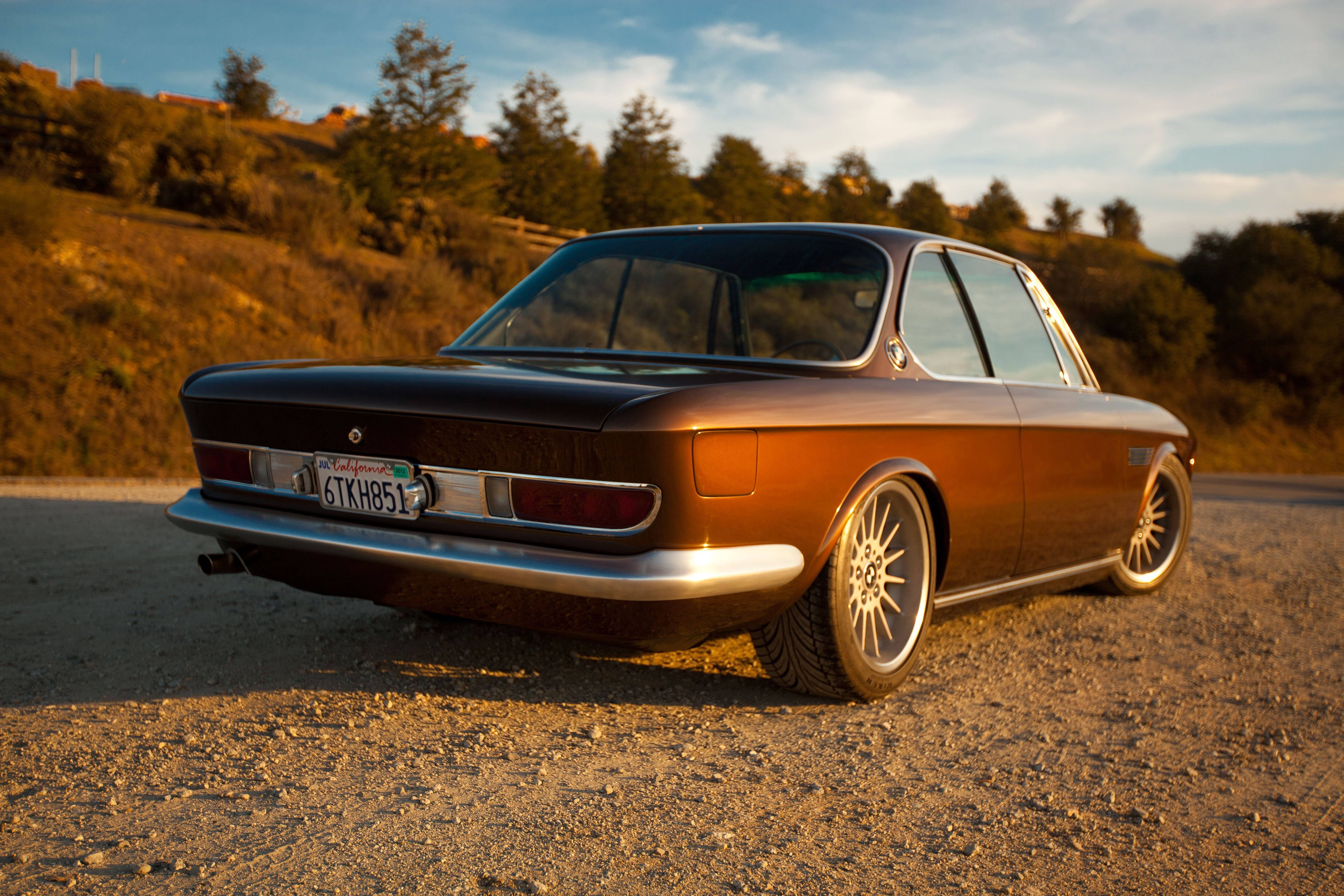 classic bmw 桌布 壁紙 — 一輛復古棕色BMW房車停泊在碎石路上，沐浴在金色夕陽中，背後山坡上有樹木。