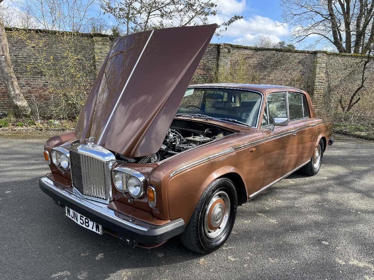 Bentley T2 桌布 壁紙 — 一輛復古棕色勞斯萊斯，引擎蓋打開，停在柏油路上，旁邊是磚牆，天空局部多雲。