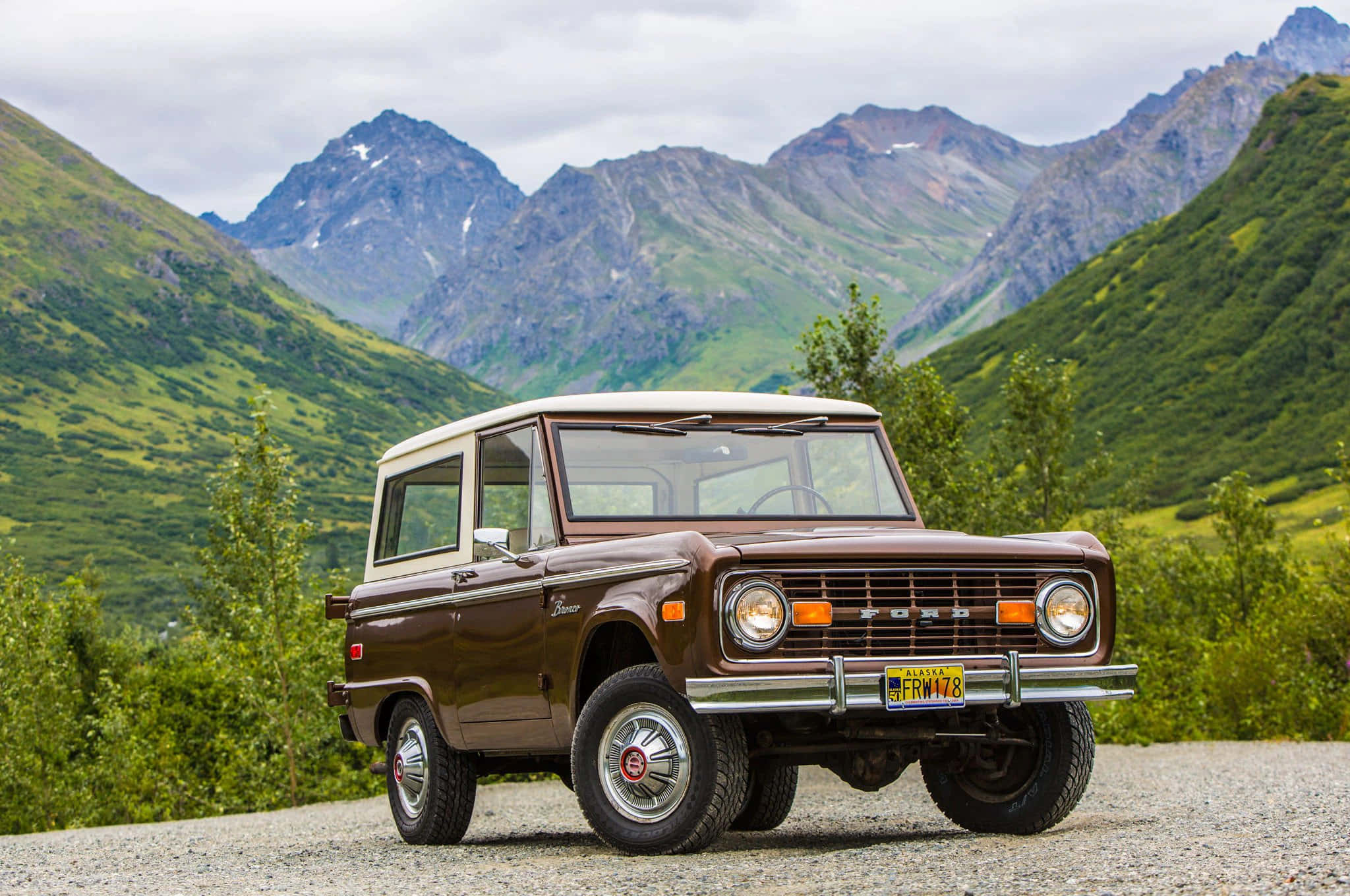 Ford Bronco 桌布 壁紙 — 一輛復古棕色福特Bronco自豪地站在碎石路上，雄偉的積雪山脈在茂密的綠色山坡後方升起。